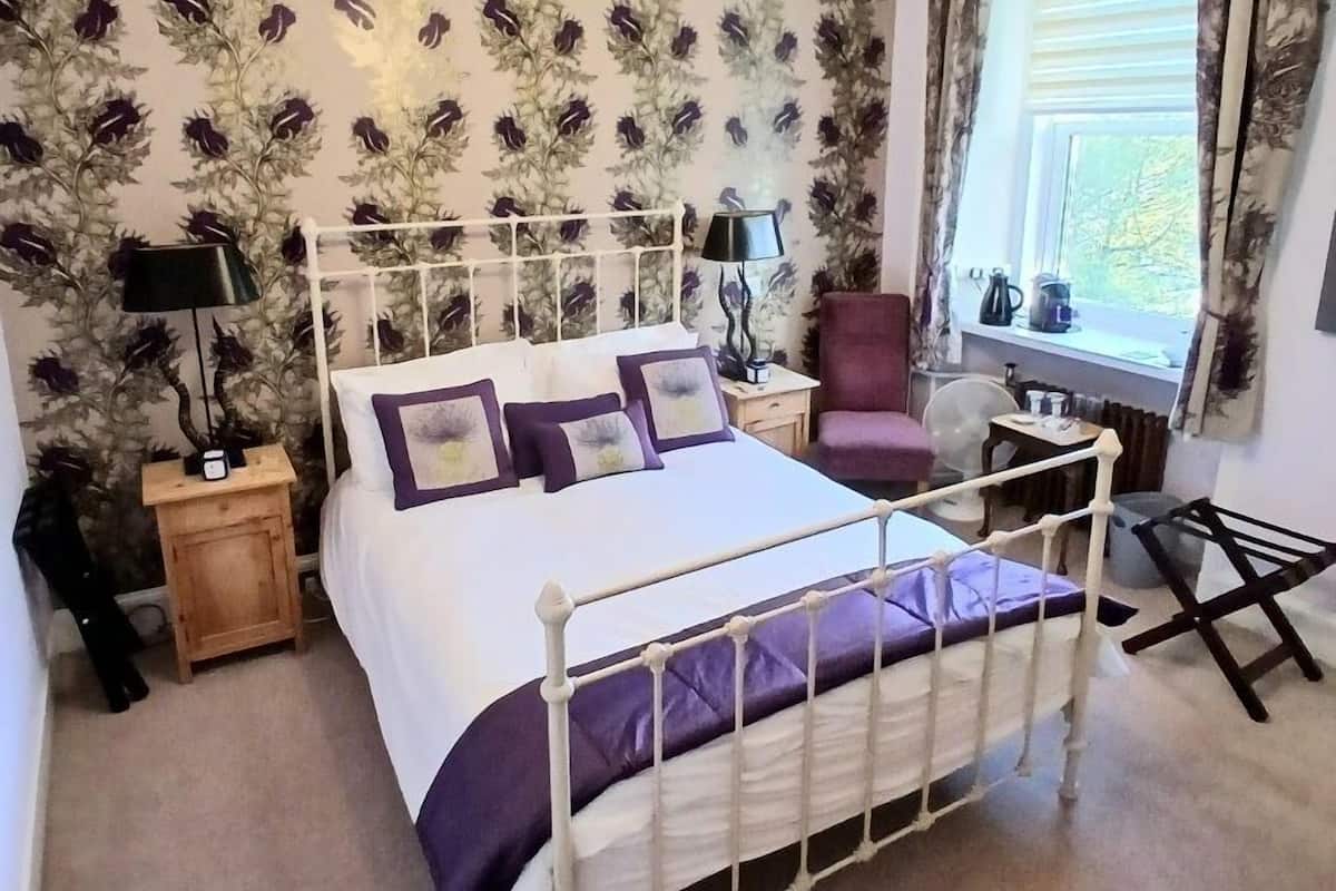 A bedroom with a bed, a nightstand, a lamp, a chair, and a window with curtains.