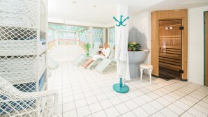Een spa ruimte met een persoon die ontspant in een stoel, een wit rek met handdoeken, een klein krukje en een houten deur.