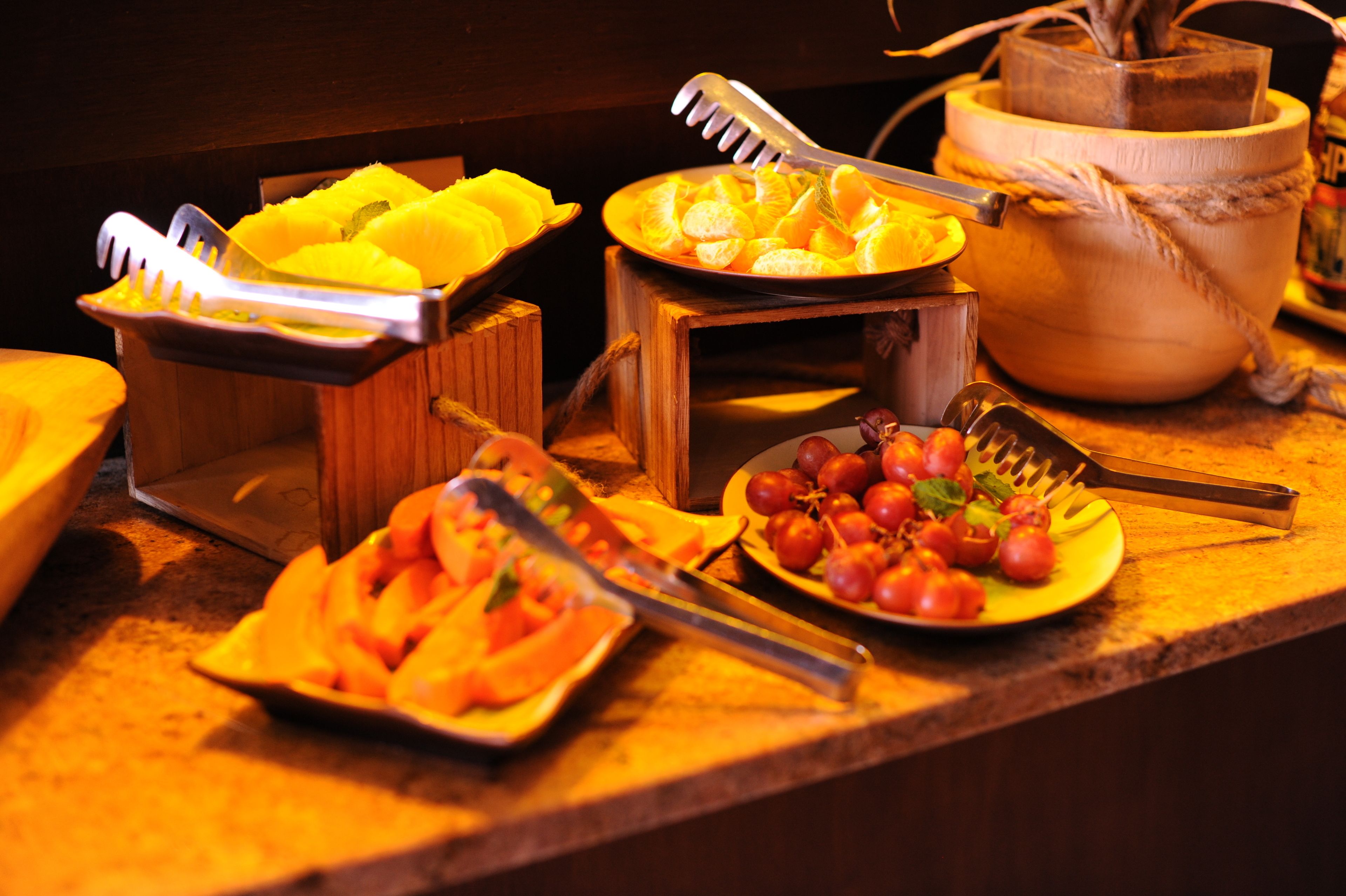 Uma mesa de buffet com diversas frutas e legumes, incluindo laranjas fatiadas, tomates e uma tigela de mangas.