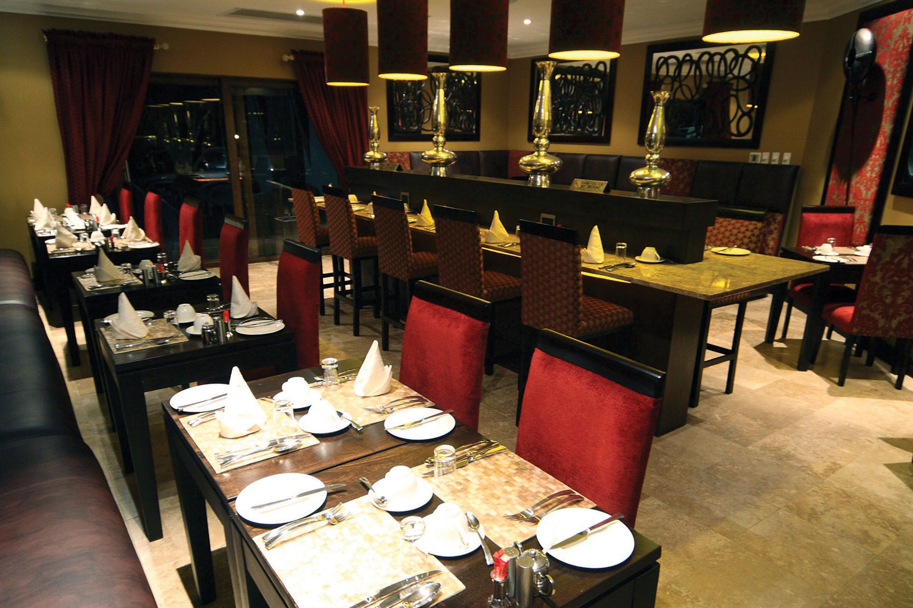 Un restaurante con varias mesas dispuestas para comer, sillas tapizadas en rojo y luminarias decorativas.