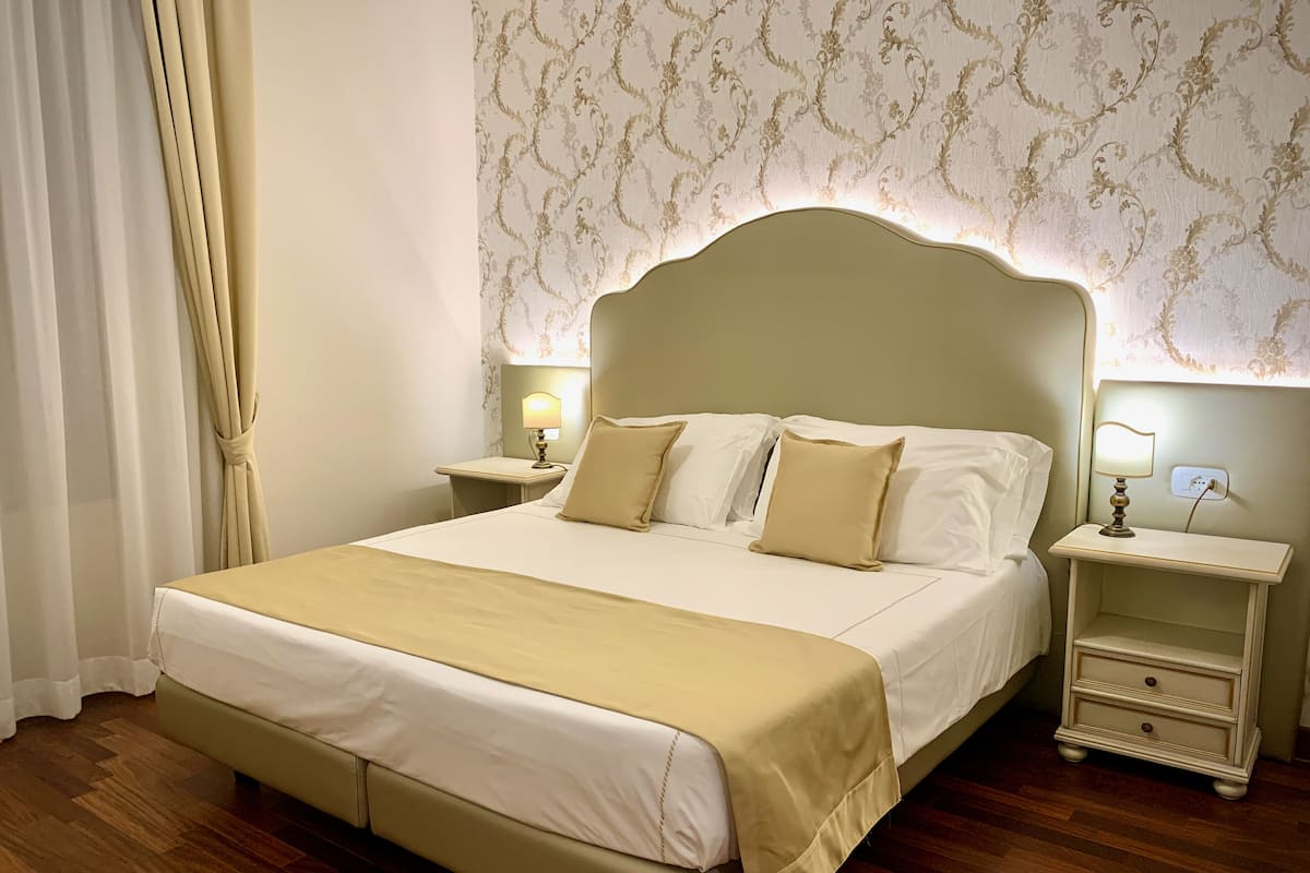 A neatly made bed with a headboard, two bedside tables with lamps, a wooden floor, and a patterned wallpaper.