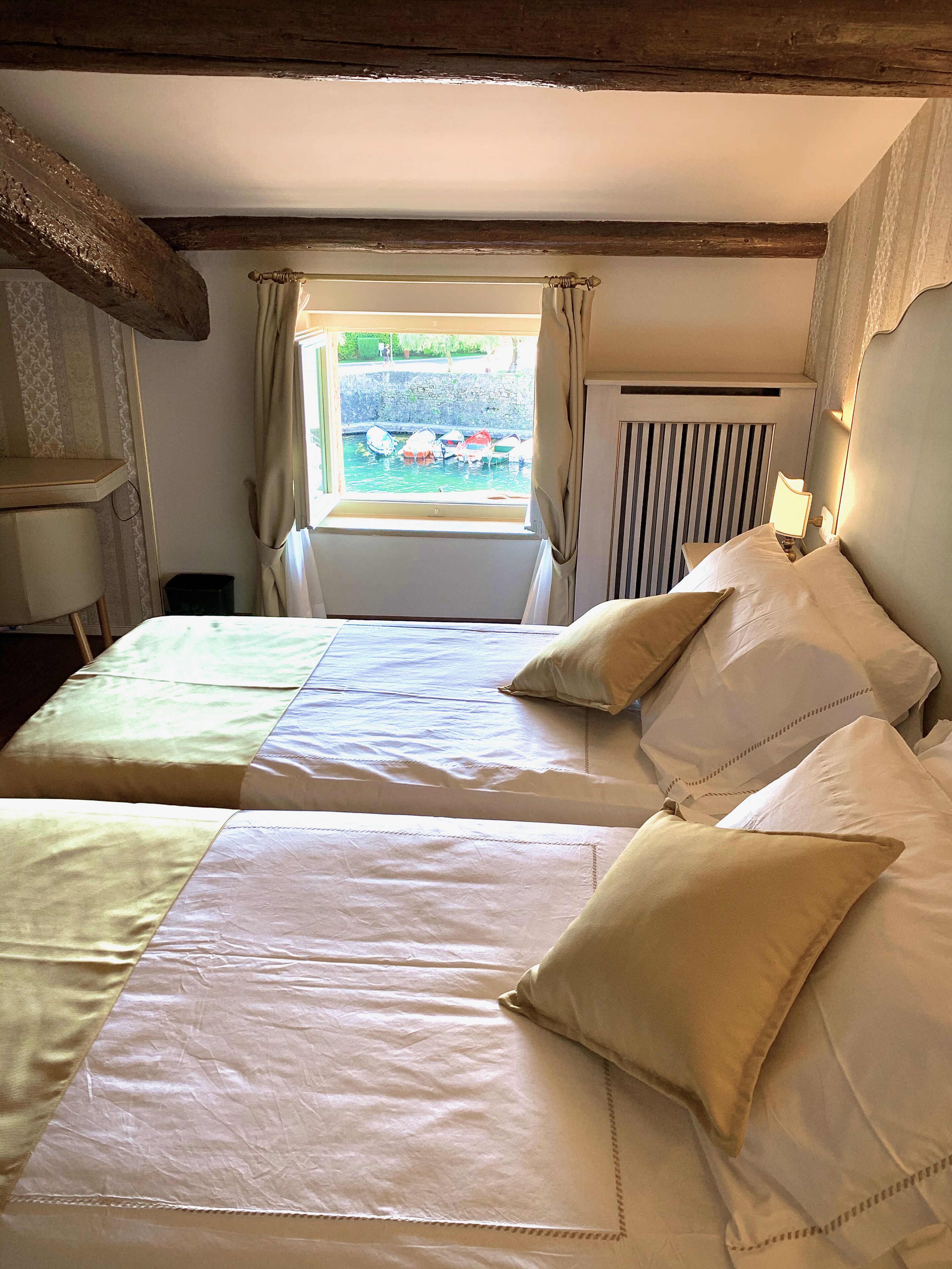 A room with two beds, a window with curtains, a radiator, and a view of boats outside.