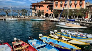 A marina with various boats docked, a building with balconies, and a clear sky. - Albergo Gardesana (Torri del Benaco)