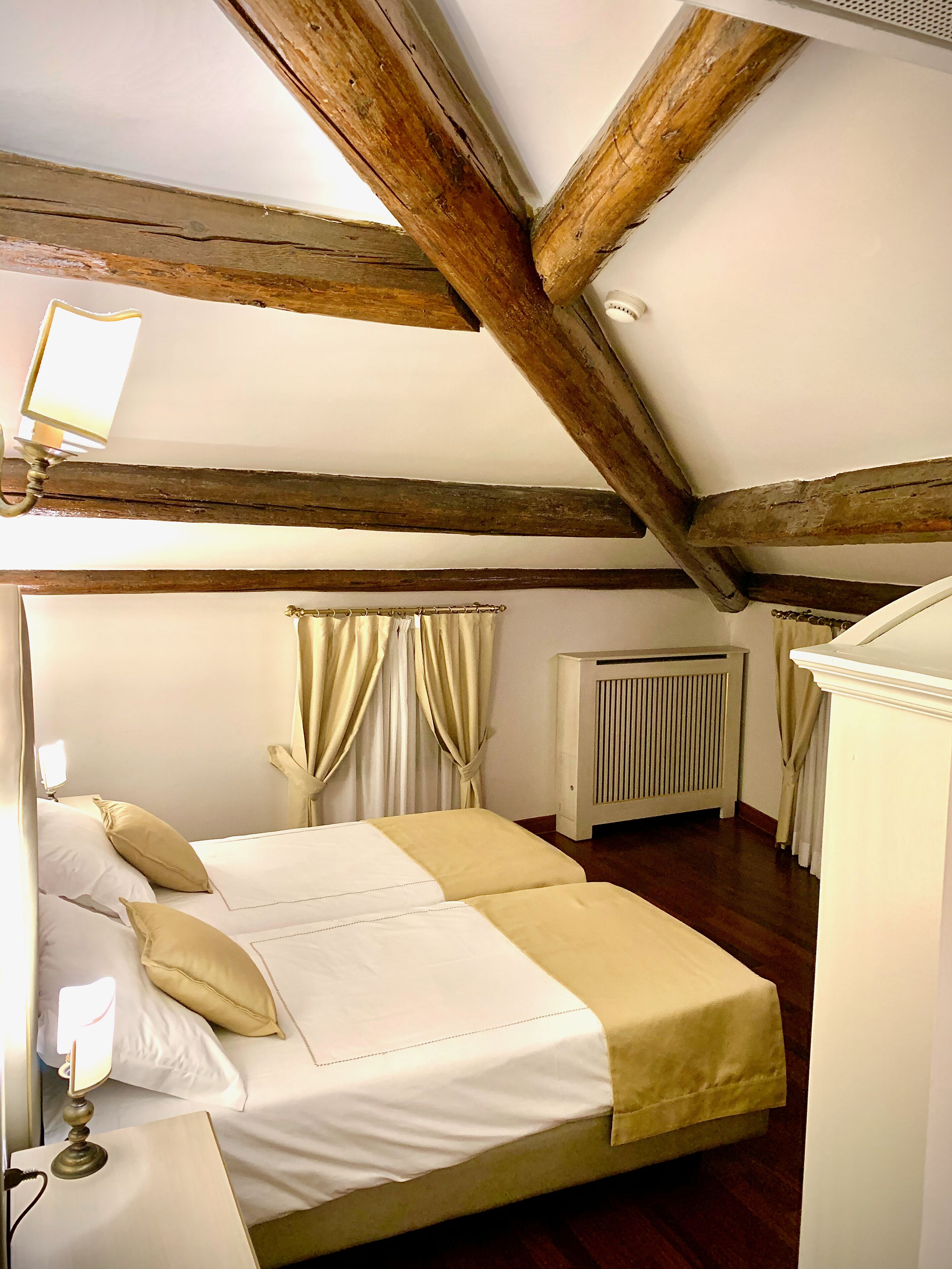 Un dormitorio con techo de madera, cama con ropa de cama blanca y beige, lámpara de noche, radiador y cortinas.