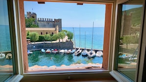 A view from a window overlooking a marina with several sailboats docked, a stone wall, and a castle-like structure in the background. - Albergo Gardesana (Torri del Benaco)