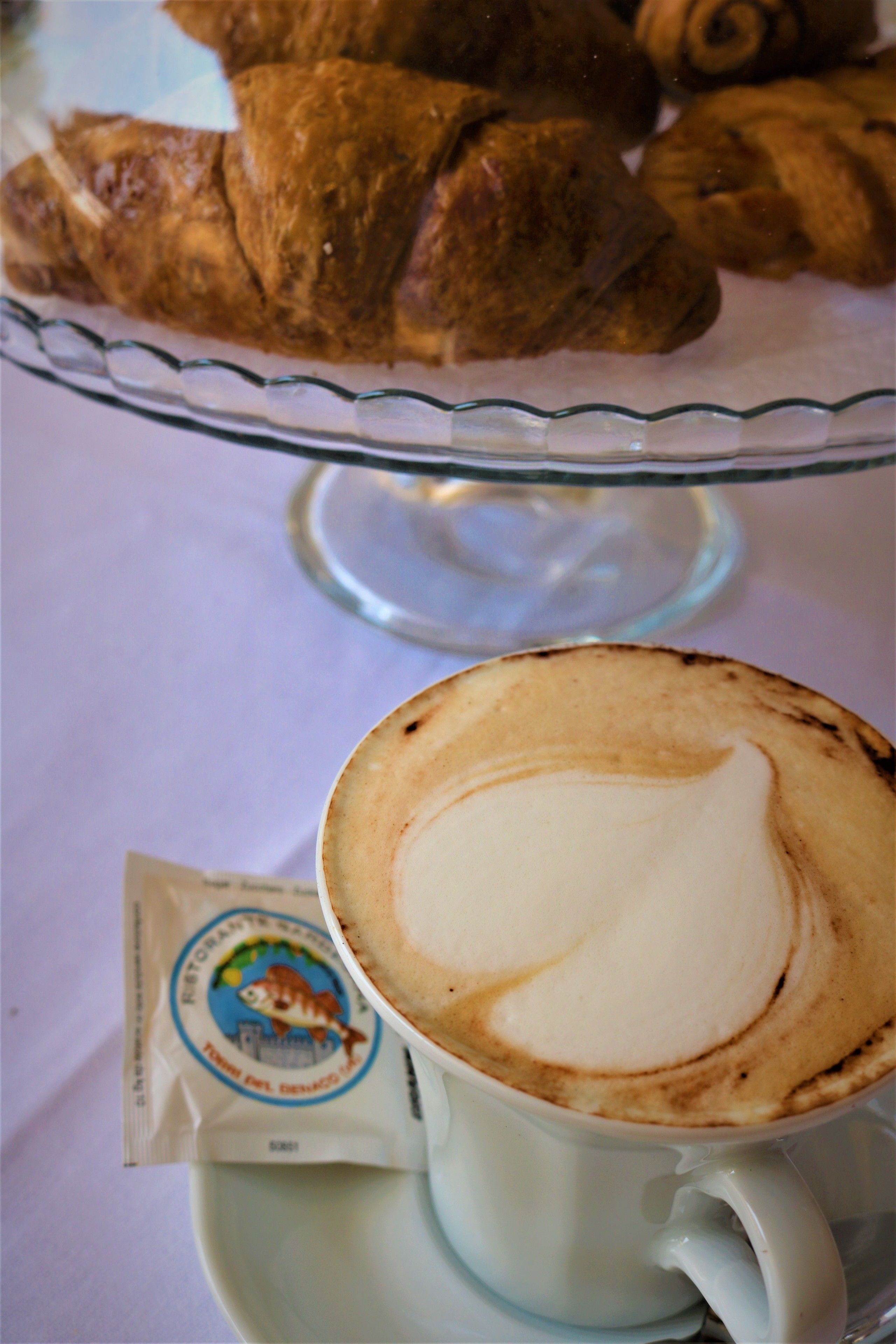 A cup of coffee with latte art on a saucer, a packet of creamer, and a glass plate with croissants.