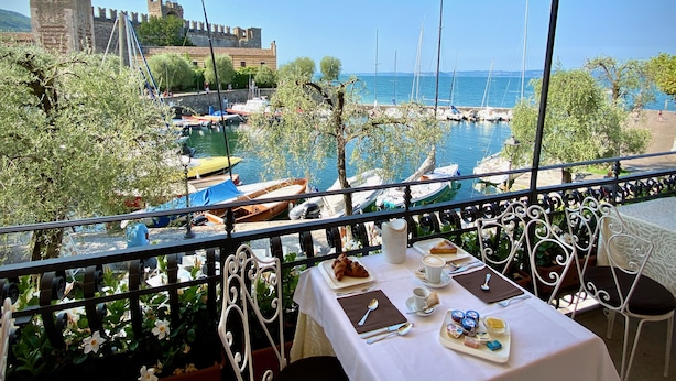 A table set with breakfast items overlooking a marina with boats and a historic building in the background. - Albergo Gardesana (Torri del Benaco)