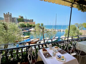 A table set with breakfast items overlooking a marina with boats and a historic building in the background. - Albergo Gardesana (Torri del Benaco)