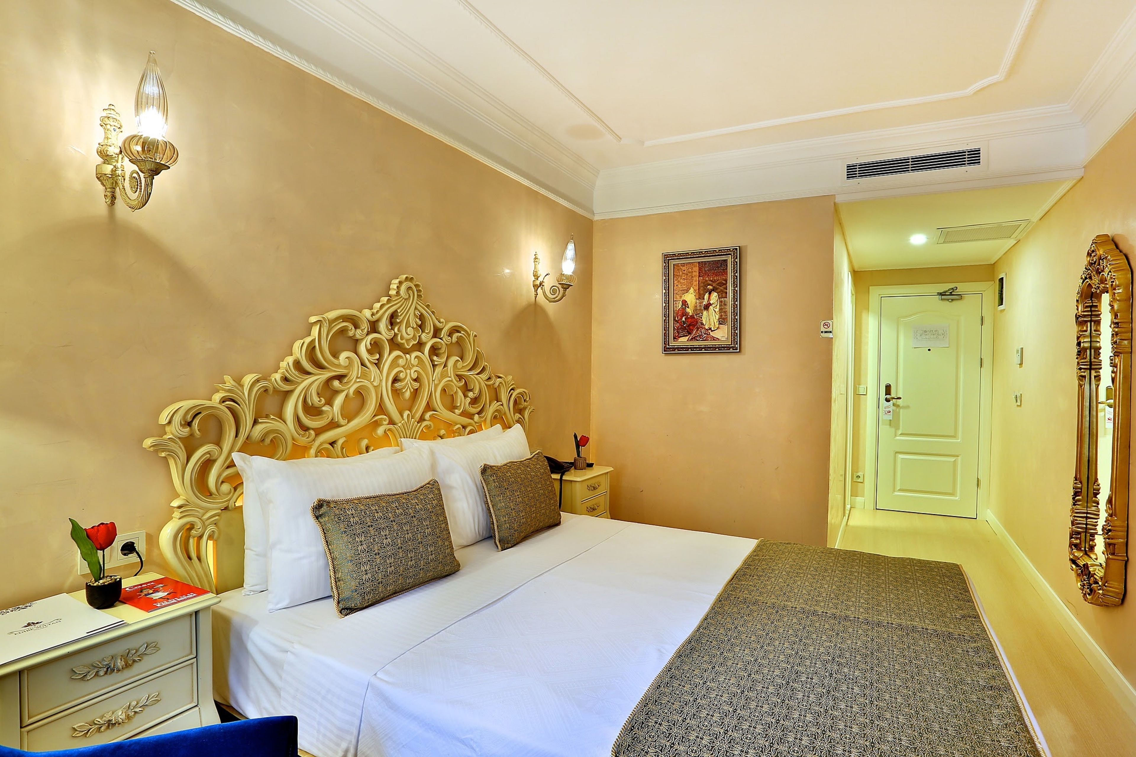 A hotel room with a large ornate bed, a nightstand with a lamp, a desk with a red book, and a mirror on the wall.