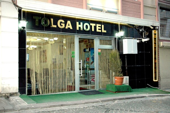 Exterior del Hotel Tolga con entrada de vidrio, felpudo verde en la puerta y una planta en maceta.