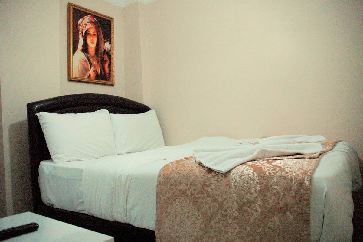 Una cama bien hecha con sábanas blancas y una manta decorativa, un control remoto sobre una mesita de noche y un cuadro en la pared.