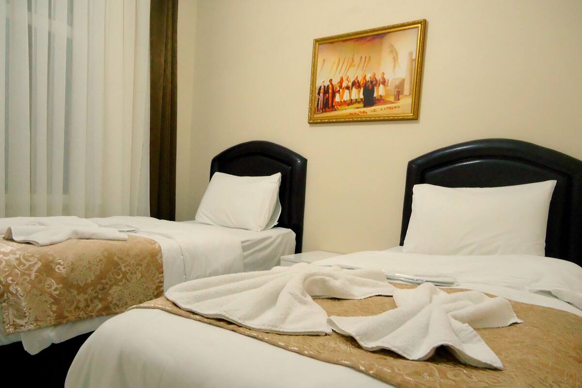 Habitación de hotel con dos camas, cada una con cabecero negro, ropa de cama blanca y colcha de cama estampada en tonos beige. Un cuadro enmarcado cuelga en la pared sobre las camas.