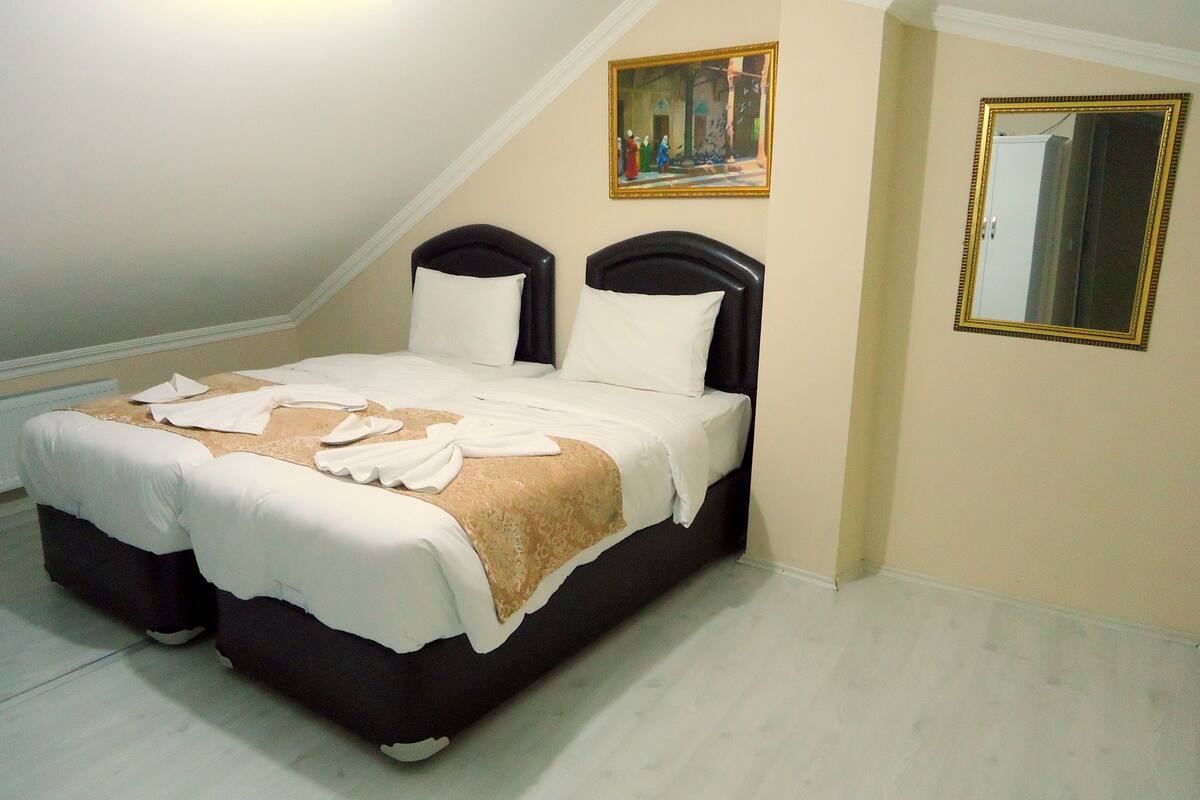 Una cama bien tendida con sábanas blancas y un plaid color beige, un cuadro encima y un espejo en la pared de al lado.