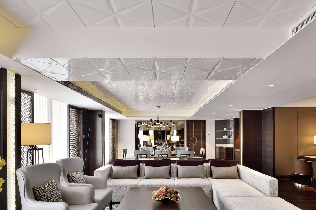 En modern hotellobby med ett geometriskt utformat tak, en sittgrupp med soffor och fåtöljer, en matplats med en stor kristallkrona och en stor spegel som reflekterar rummet.