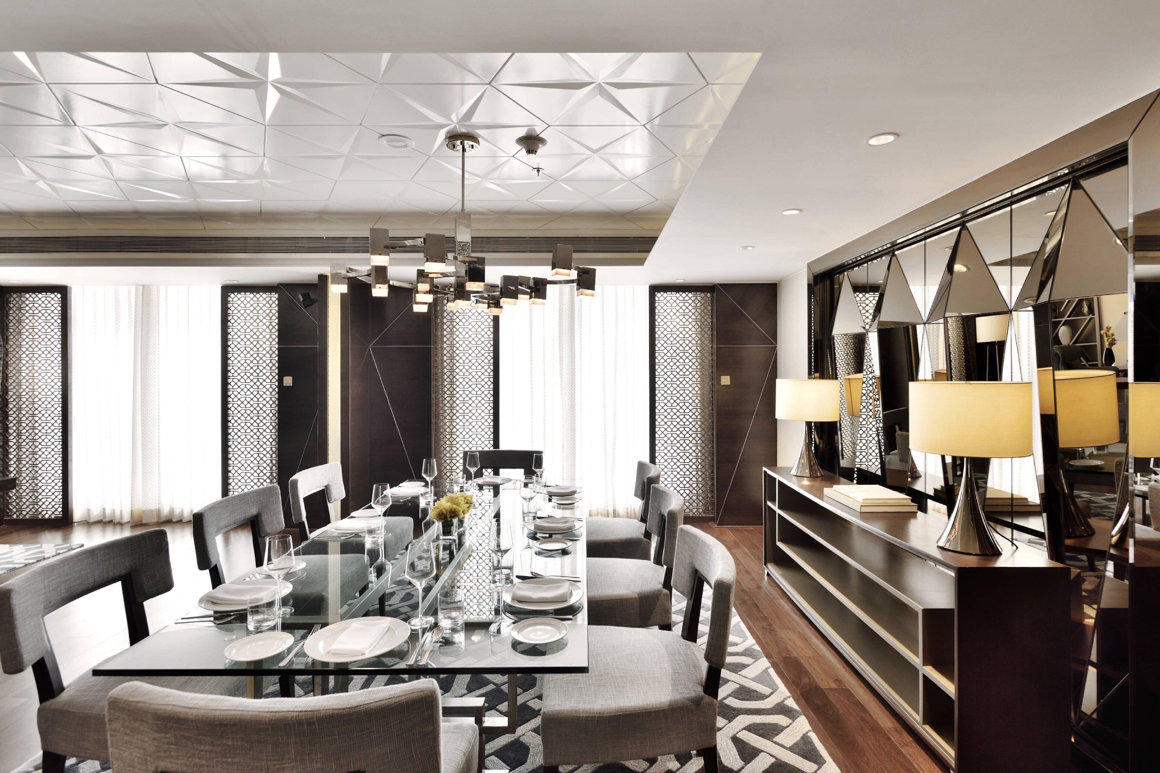 A modern dining area with a glass table, grey chairs, and a geometric ceiling design.