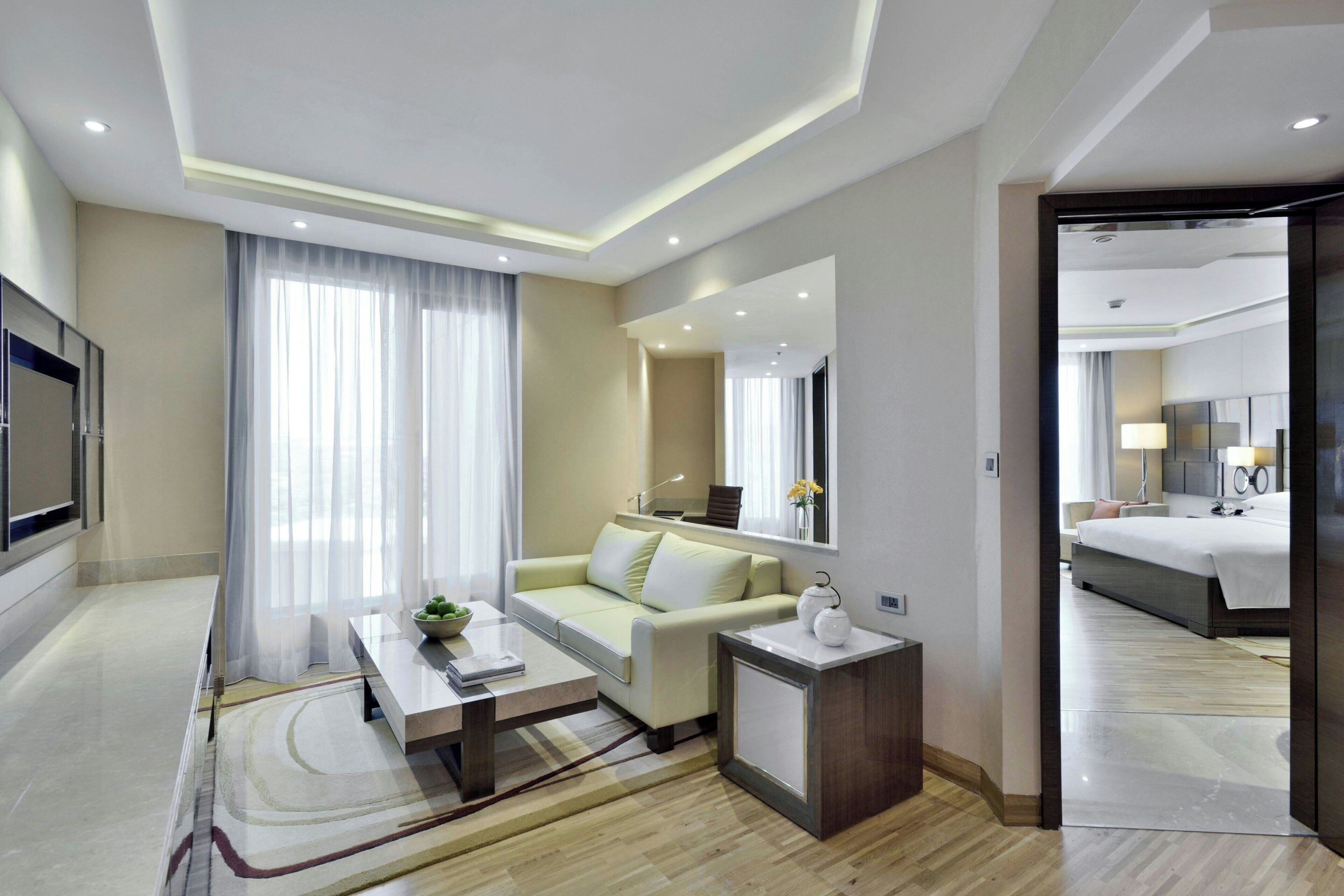 Ein modernes Hotelzimmer mit einer Couch, einem kleinen Tisch, einem Nachttisch, einem Spiegel und Blick in das angrenzende Schlafzimmer.