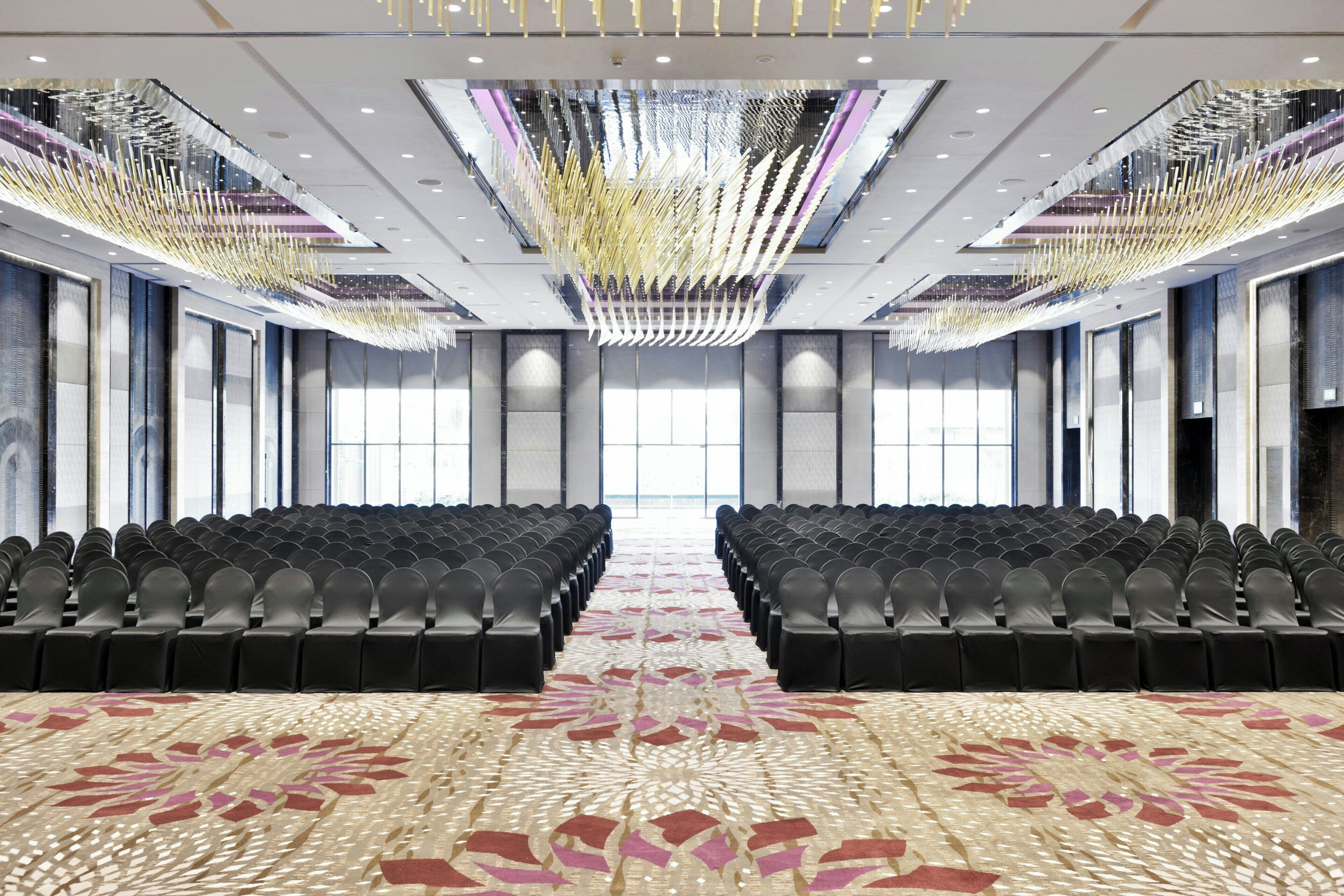 Una amplia sala de conferencias con filas de sillas, una alfombra estampada y una lámpara de araña moderna.