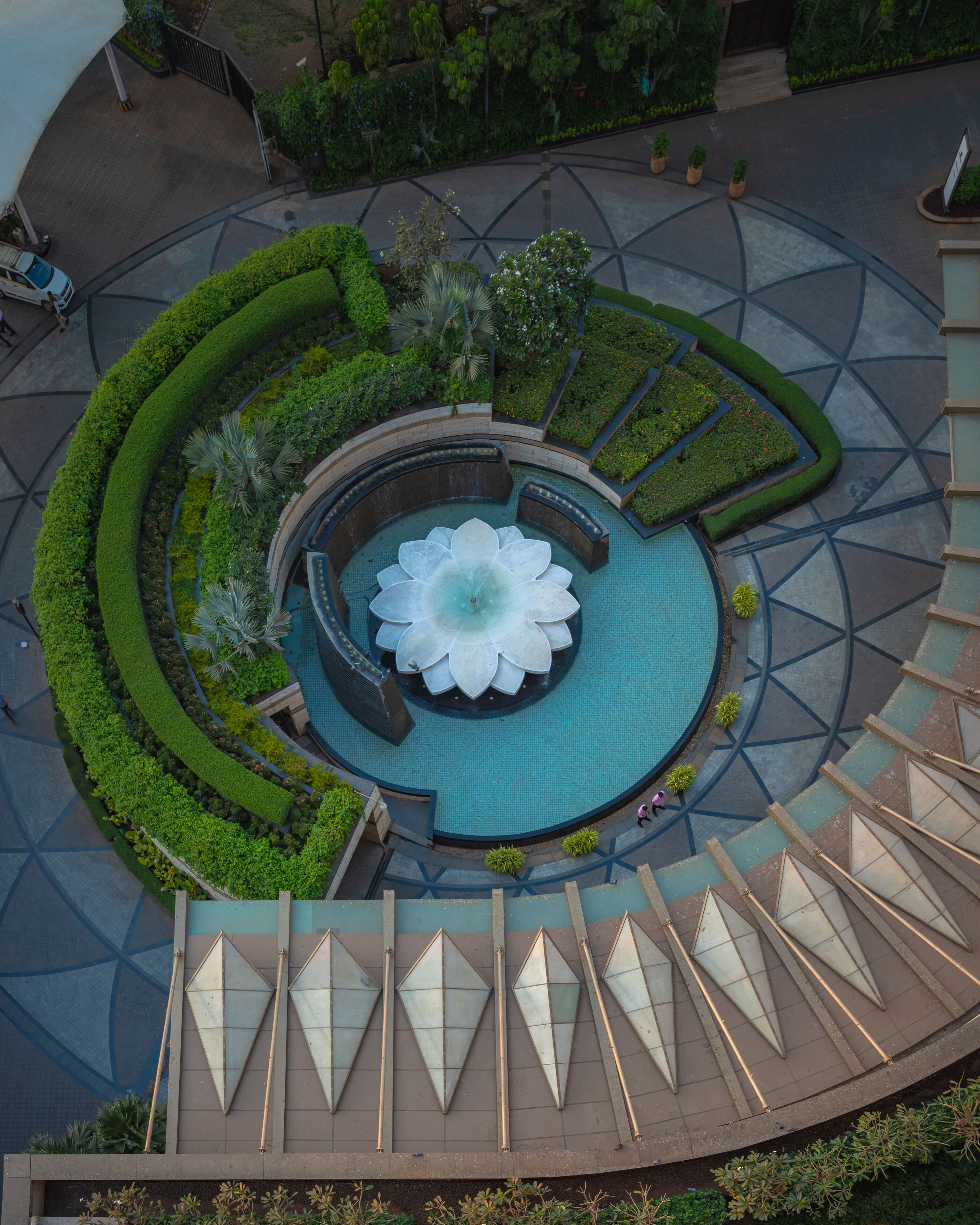 Uma fonte circular com design de lótus, rodeada por um jardim ajardinado com padrões geométricos e uma estrutura arquitetônica moderna com elementos triangulares.
