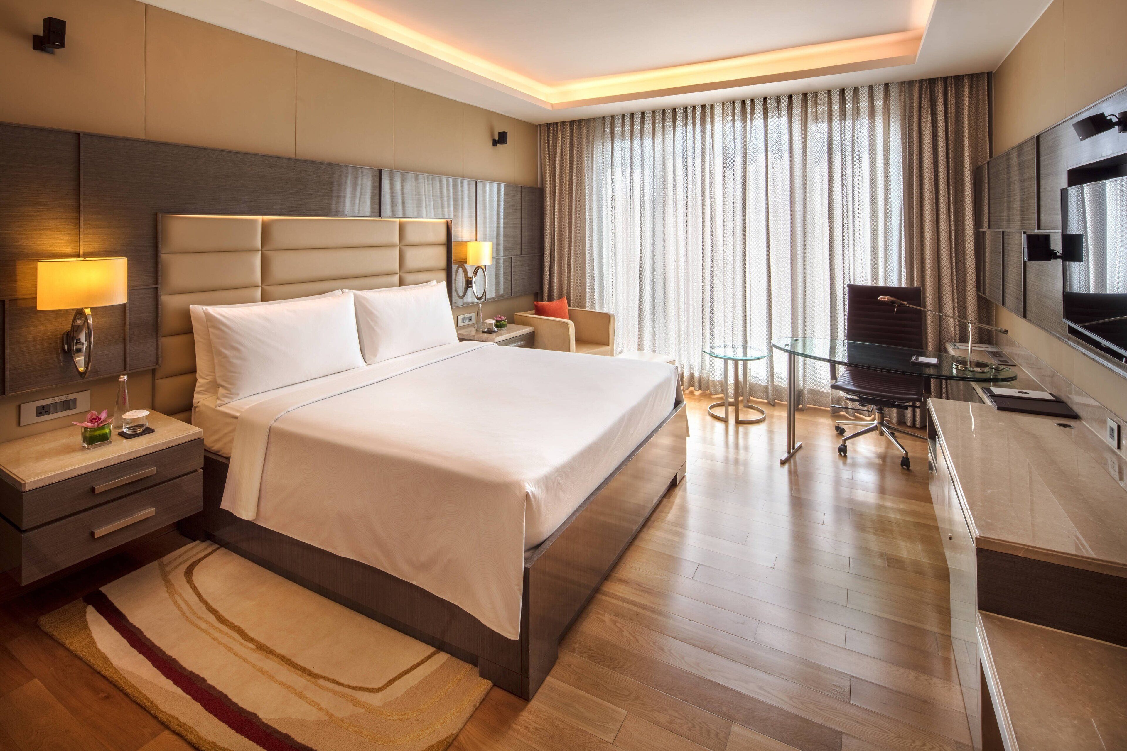 Quarto de hotel moderno com uma cama grande, uma escrivaninha com tampo de vidro, uma televisão e um espaço de estar com uma cadeira e mesa.