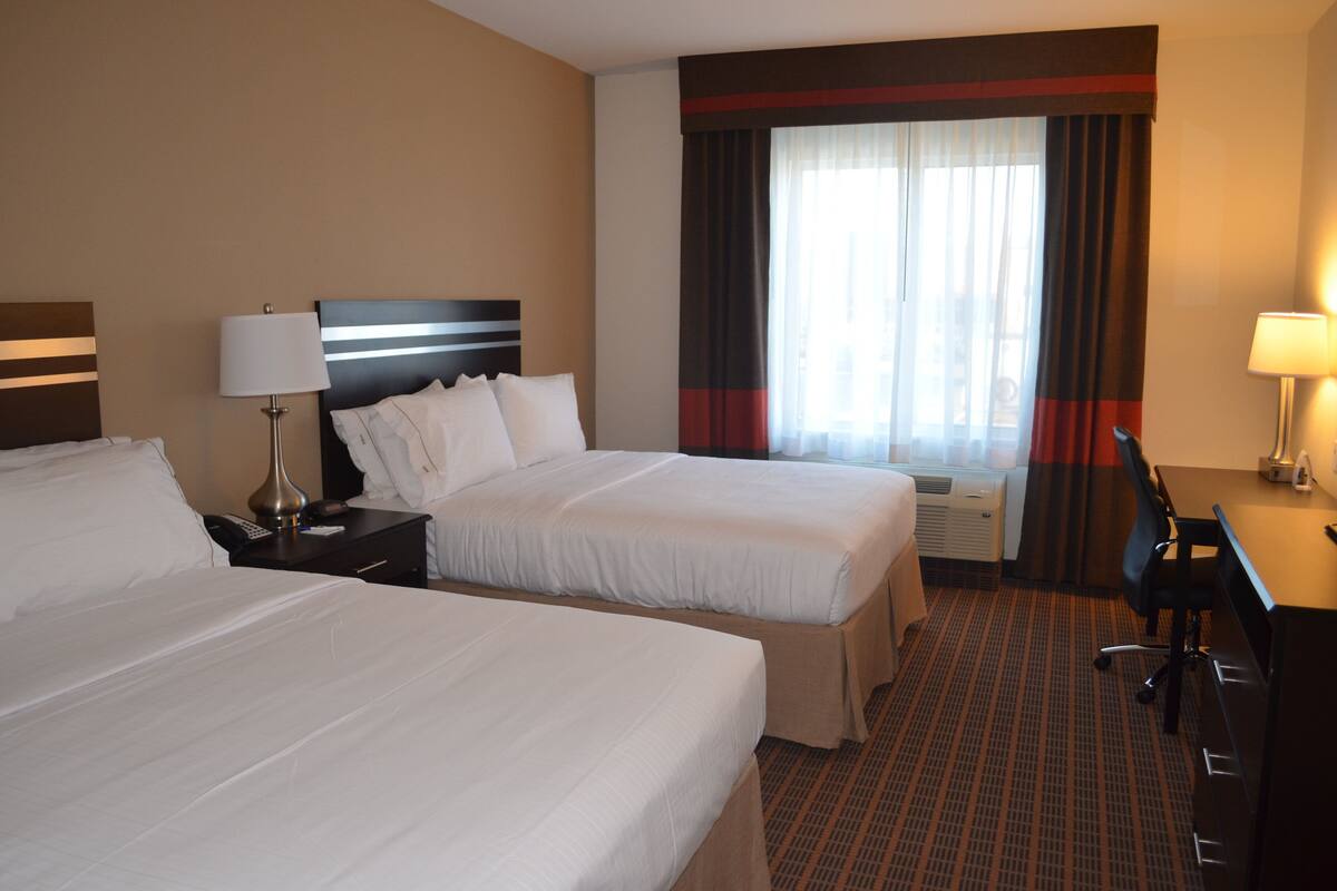 A hotel room with two beds, a desk, a chair, a lamp, and a window with curtains.