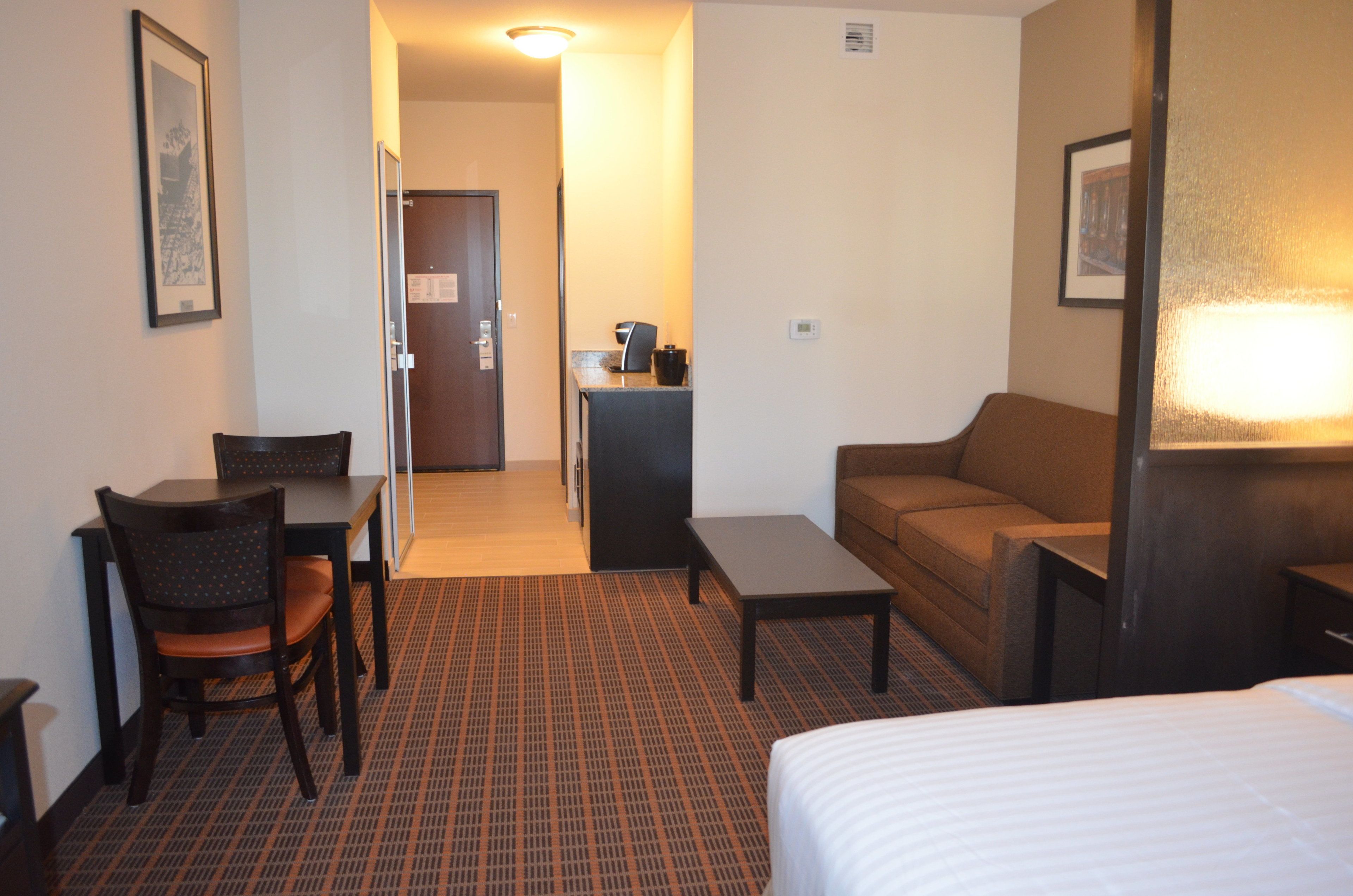 Une chambre d’hôtel comprenant un lit, un canapé, une table à manger avec des chaises, une petite table avec une lampe et une porte donnant sur un couloir.
