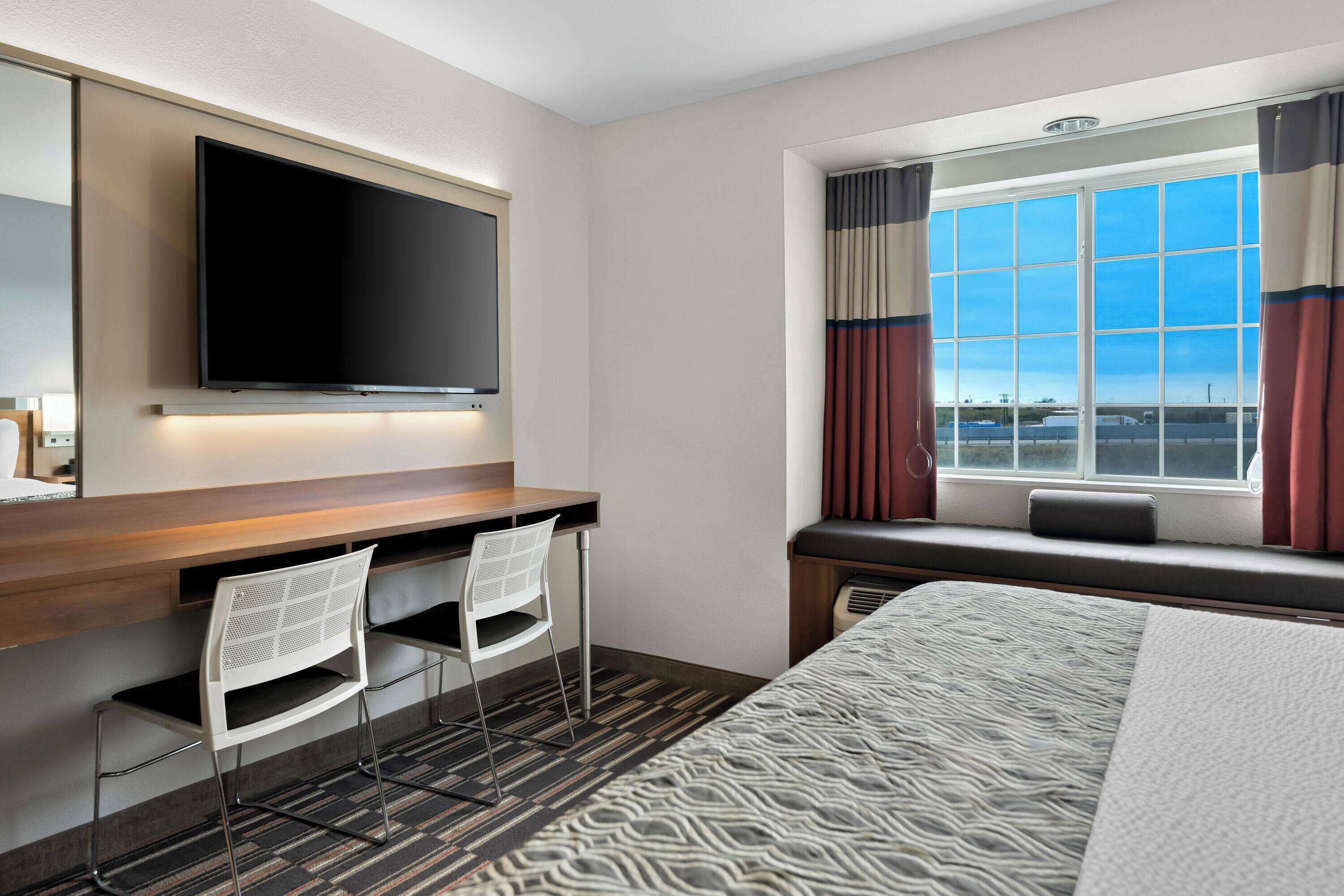 Habitación de hotel con una cama grande, un escritorio con dos sillas, un televisor de pantalla plana en la pared y una ventana con cortinas.