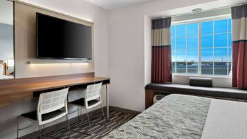 Habitación de hotel con una cama grande, un escritorio con dos sillas, un televisor de pantalla plana en la pared y una ventana con cortinas.