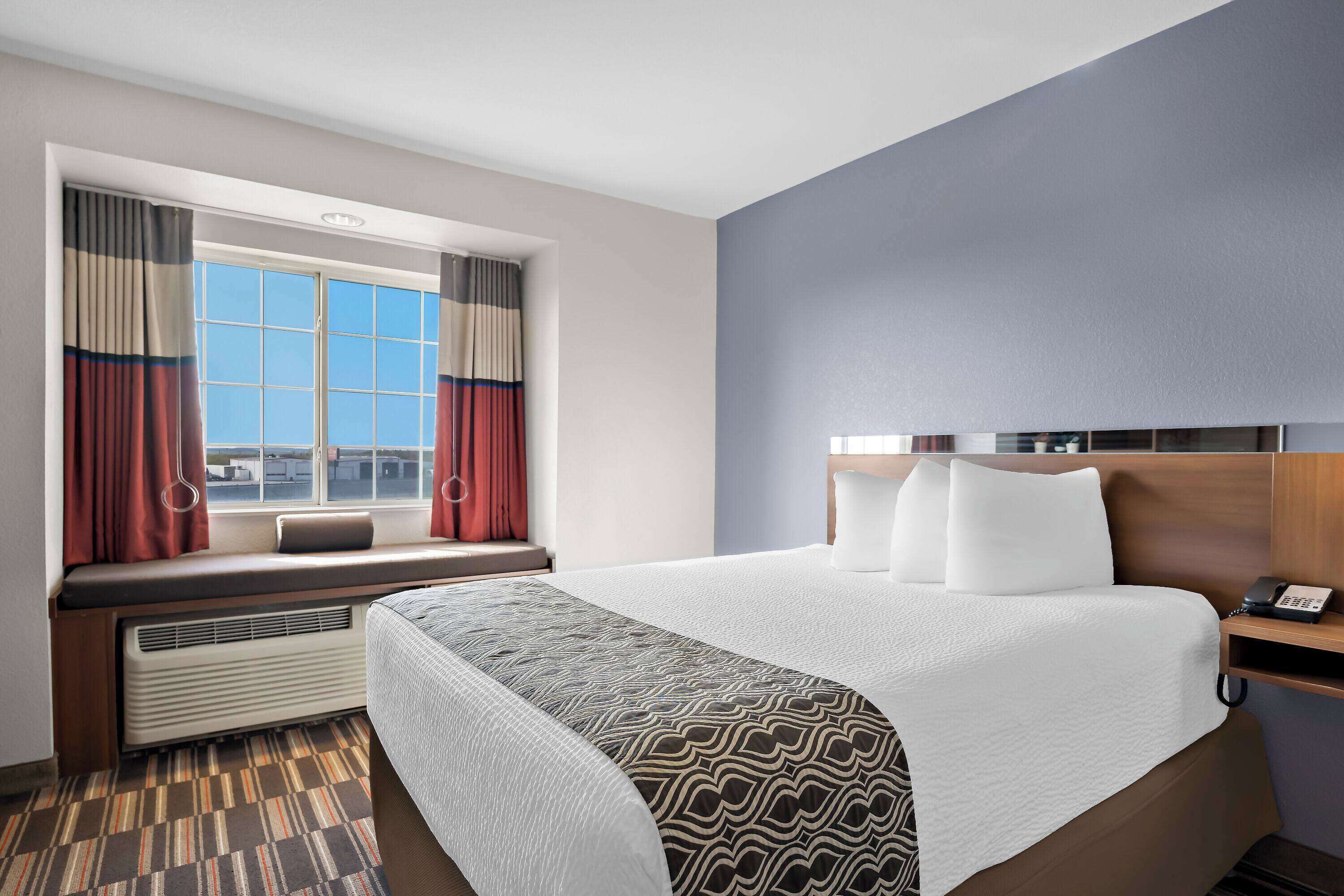 Ein Hotelzimmer mit einem großen Bett, einem Fenster mit Vorhängen, einem Nachttisch mit Telefon und einer fest installierten Heizung.
