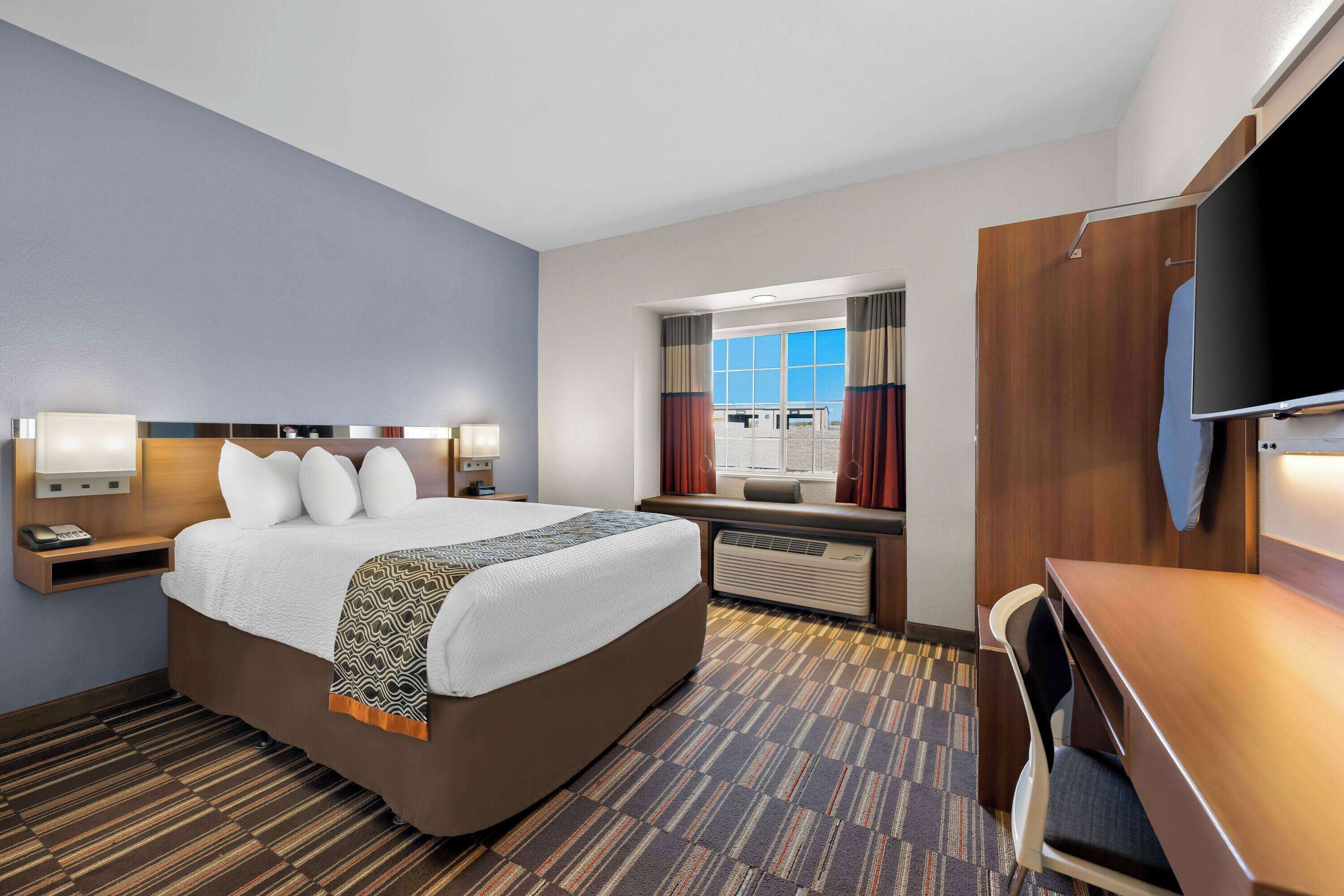 A hotel room with a large bed, bedside tables with lamps, a desk with a chair, a TV, and a window with curtains.