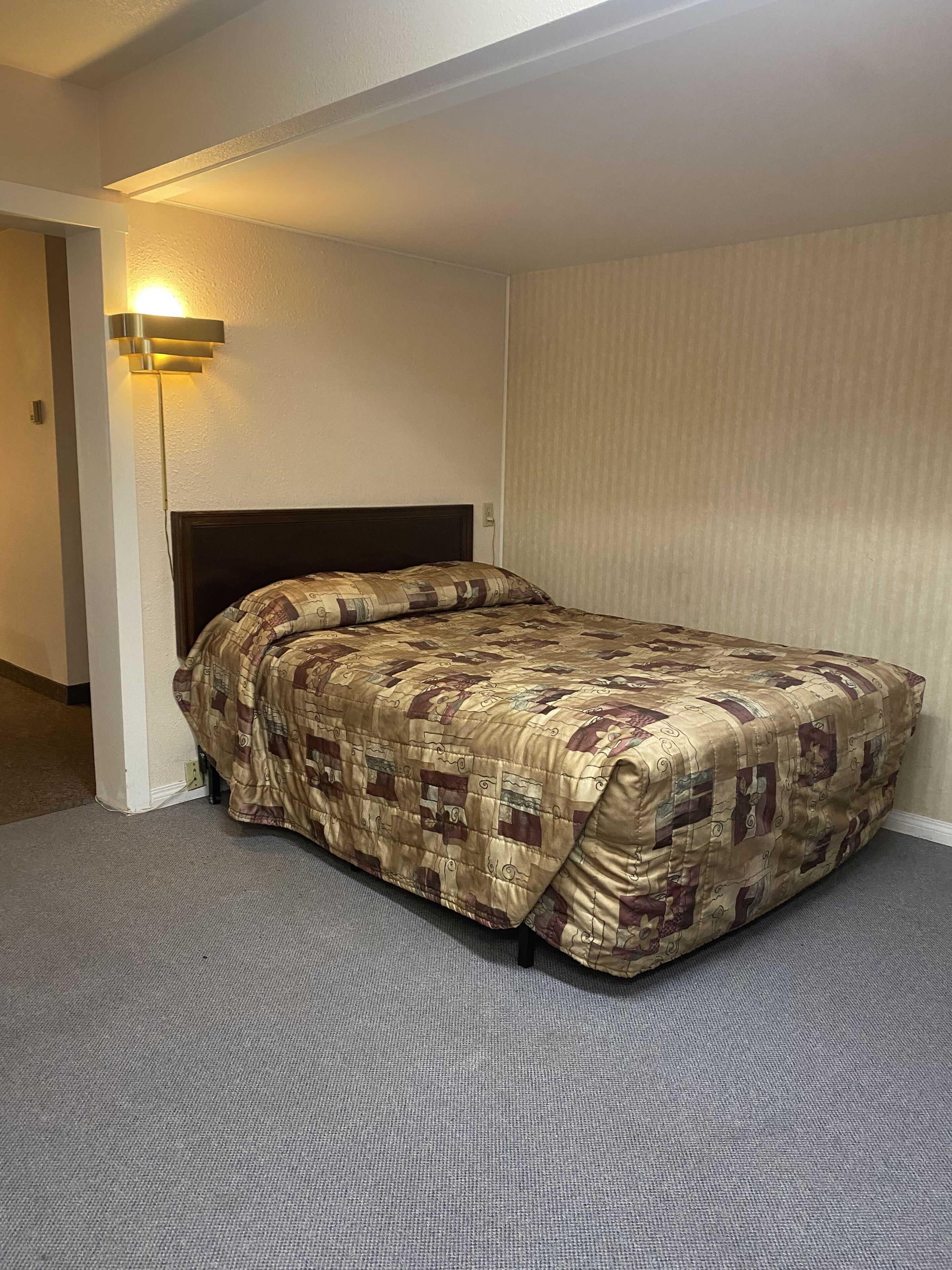 A single bed with a quilt in a room with a carpeted floor and a wall-mounted light fixture.