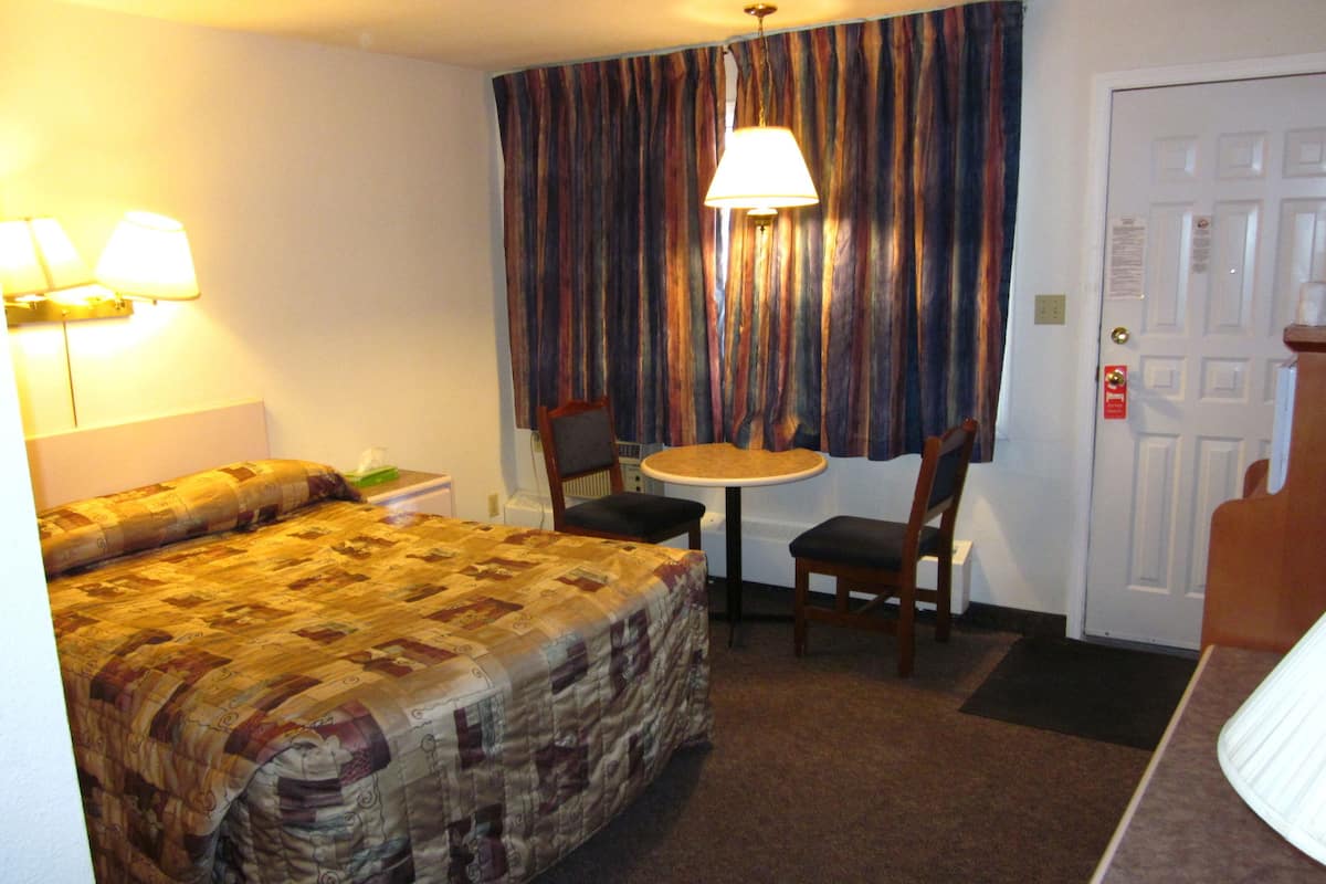 Quarto de hotel com cama, uma mesa pequena, duas cadeiras, um abajur e uma porta.