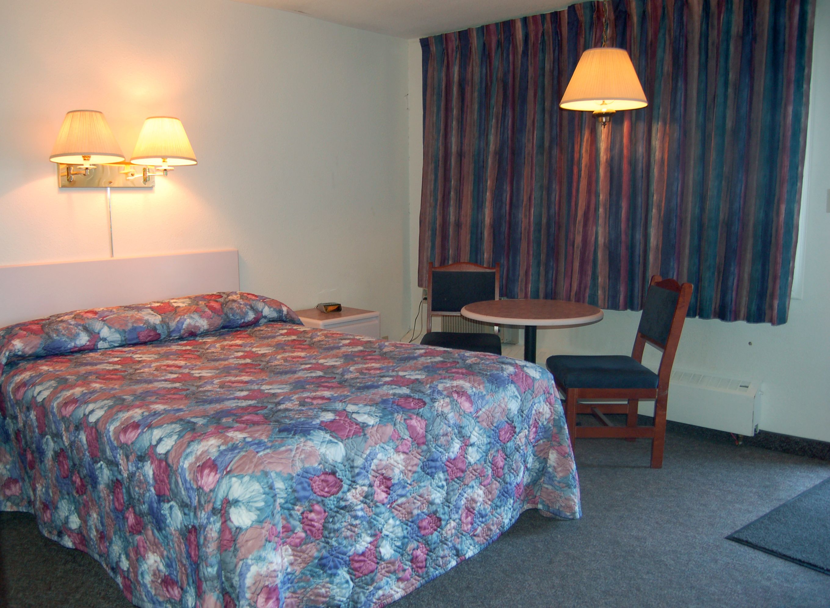 A hotel room with a bed, two wall-mounted lamps, a small table with a chair, and a window with curtains.