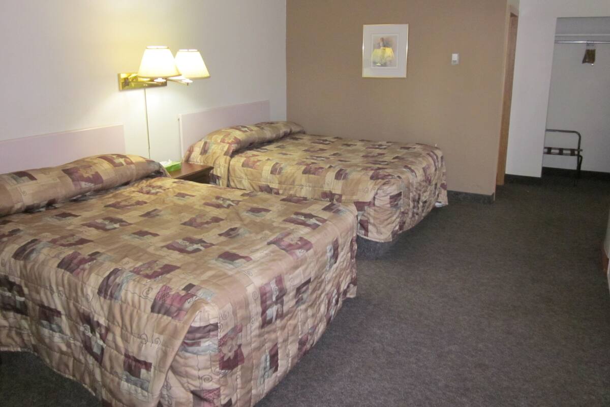Quarto de hotel com duas camas, uma mesinha de cabeceira, uma luminária e um quadro na parede.