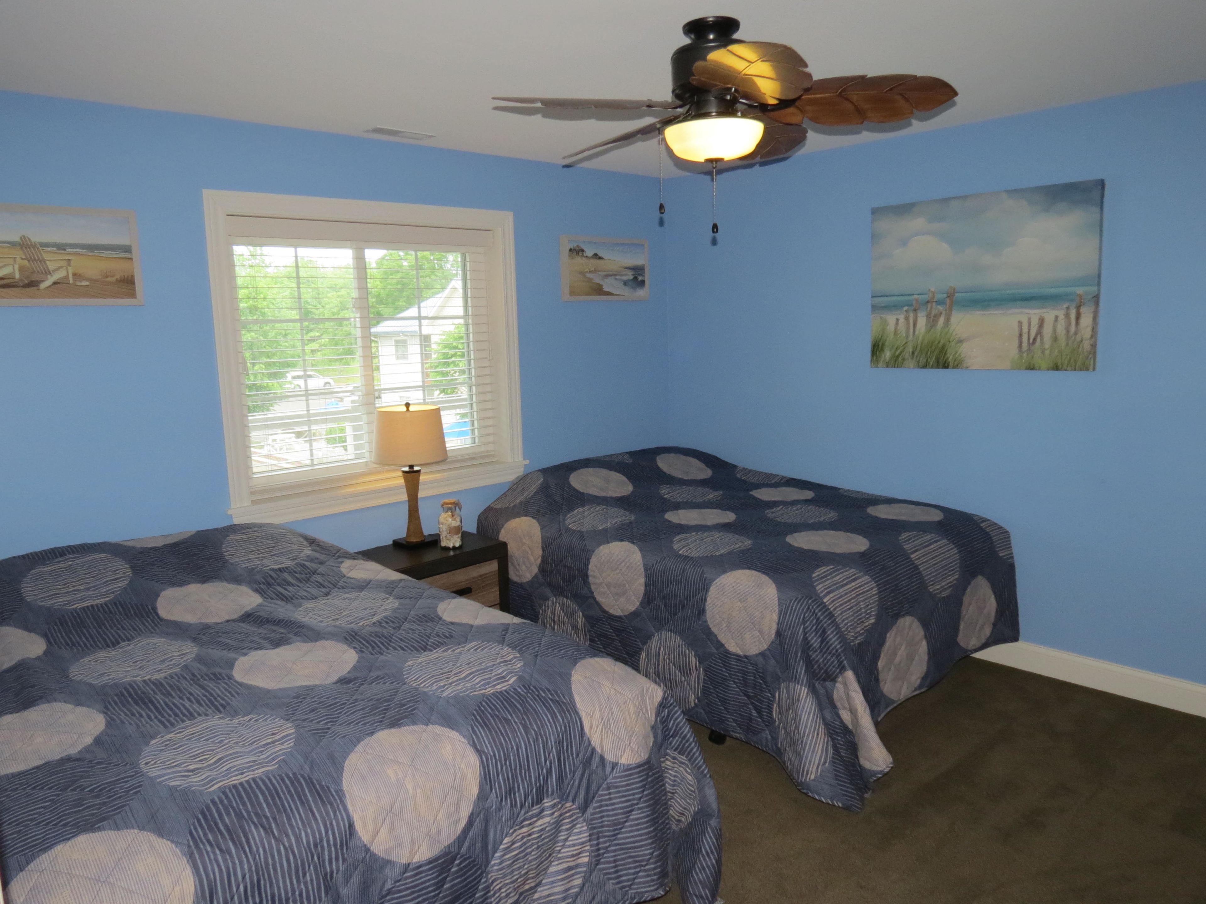 A bedroom with two beds, a ceiling fan, a window with blinds, and beach-themed artwork on the walls.
