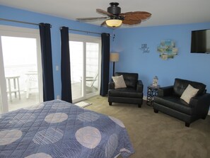 A bedroom with a bed, two armchairs, a ceiling fan, and a view to the outside.