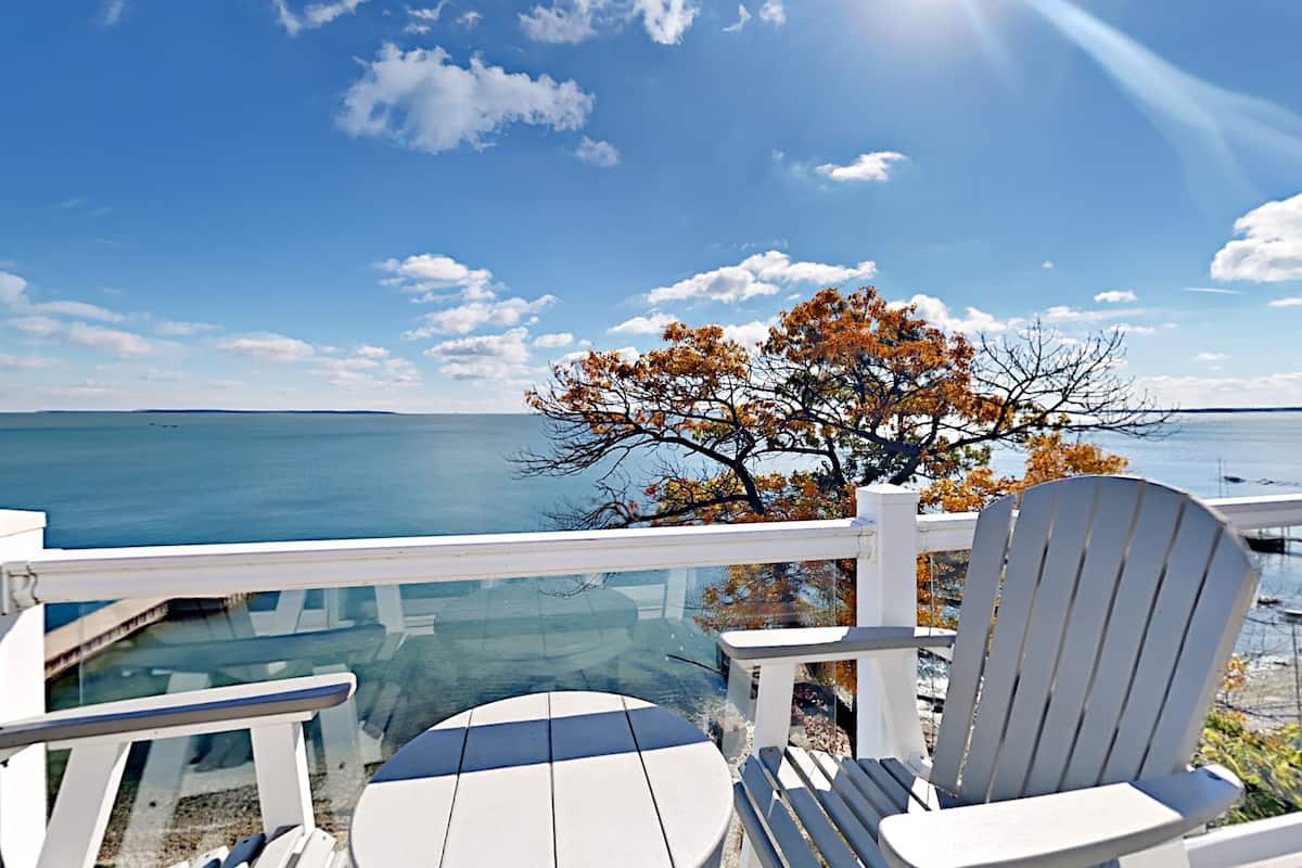 Um terraço com cadeiras Adirondack brancas e uma mesa de vidro, com vista para um corpo d'água e uma árvore com folhas outonais.