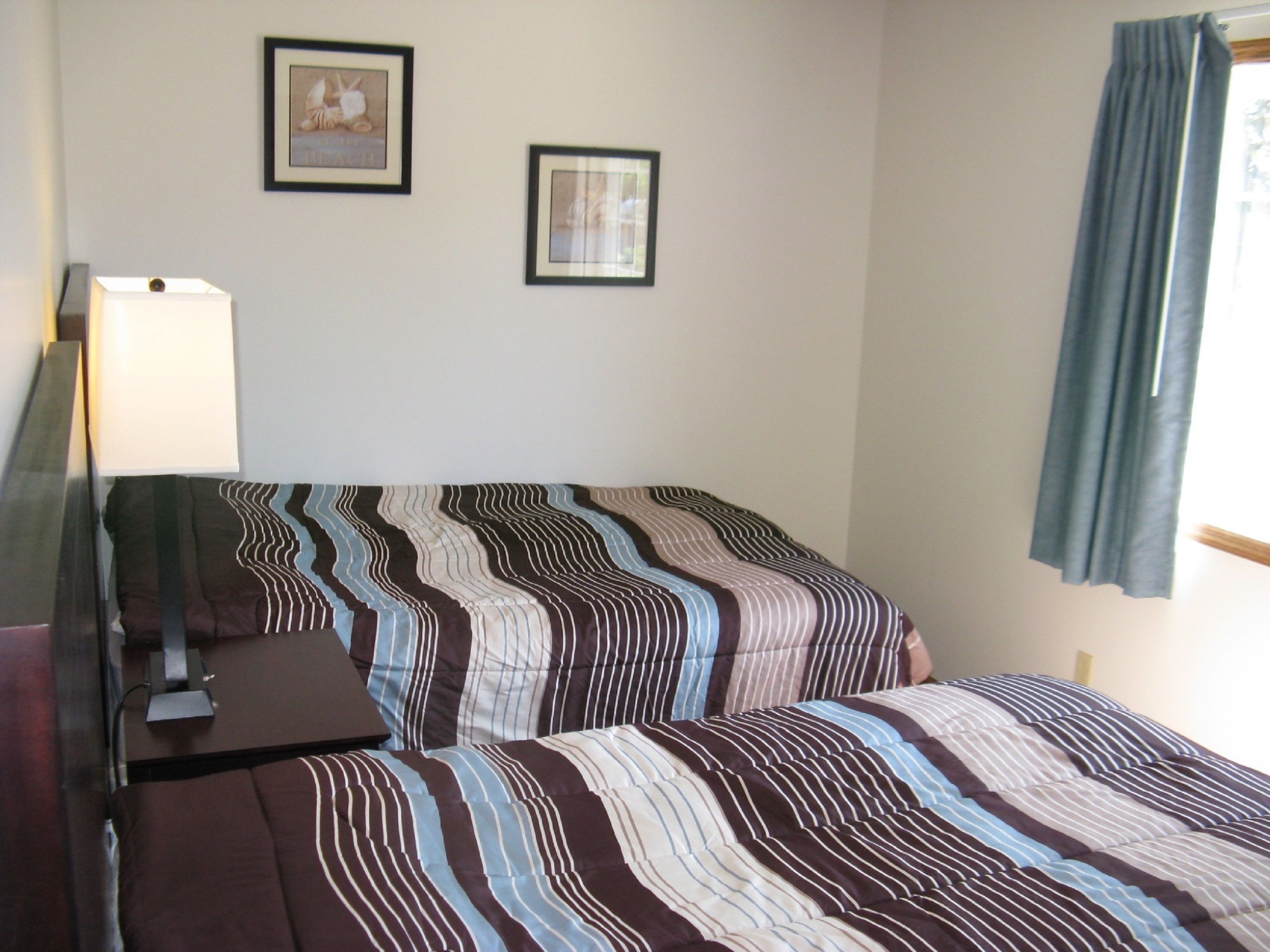 A hotel room with a bed, a desk, and a lamp. There are two framed pictures on the wall.