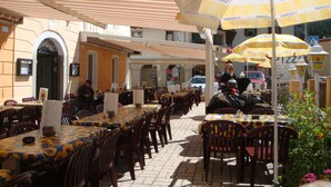 Un café al aire libre con mesas y sillas, cubierto por sombrillas a rayas, y un edificio con un letrero que dice "CAFFE".