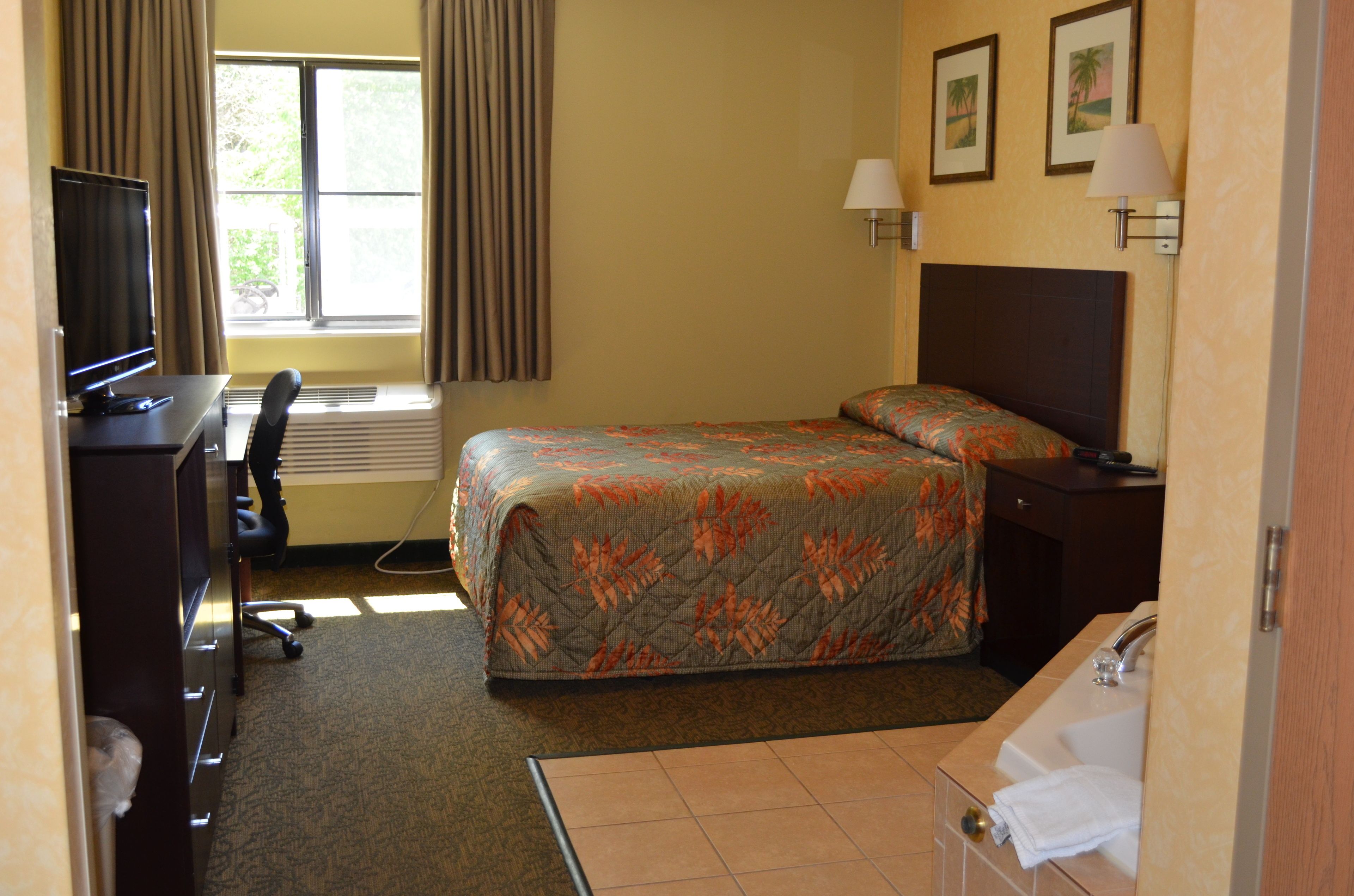 Habitación de hotel con cama, escritorio con computadora, silla, ventana con cortinas, lámpara de pared y baño con lavamanos y toalla.