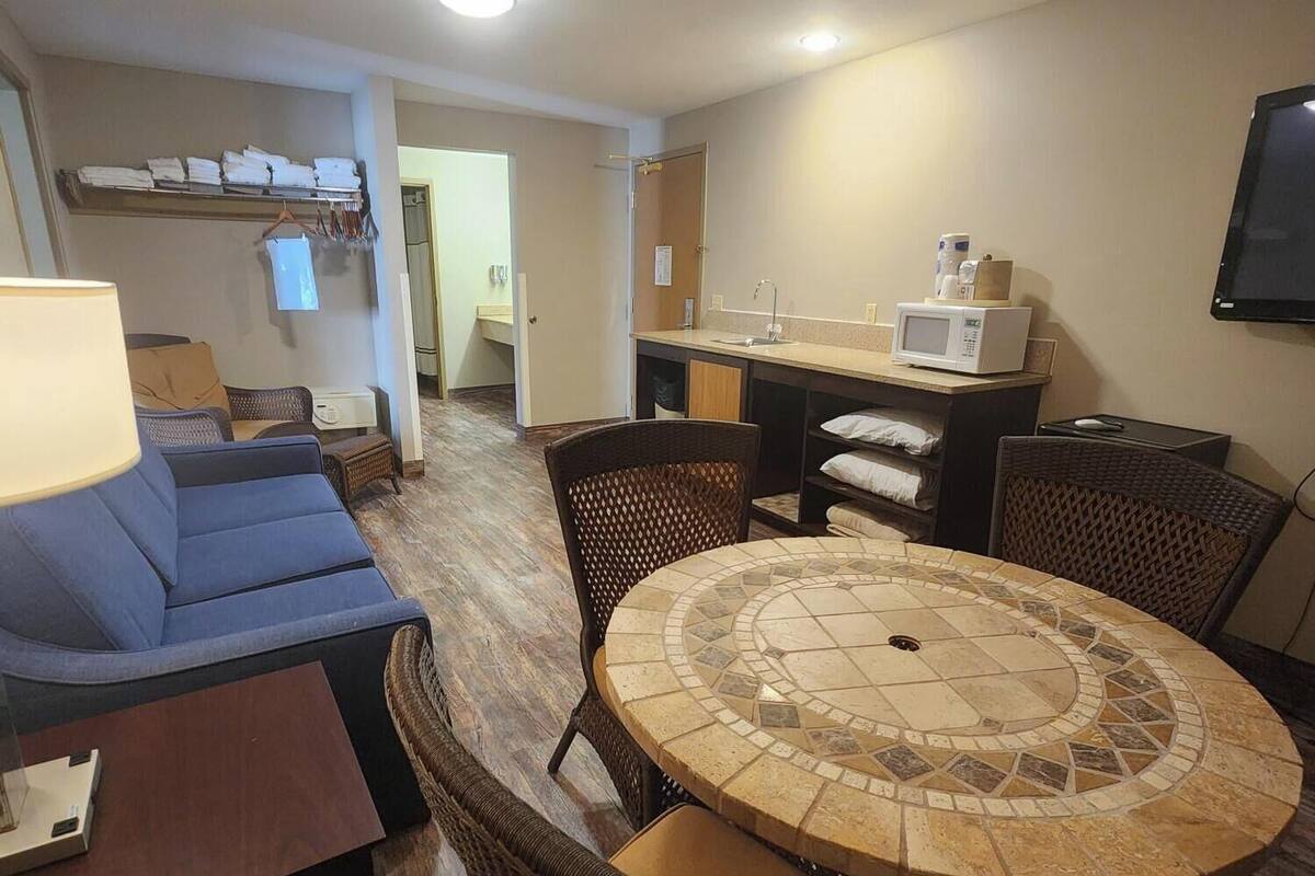 A compact living space with a sofa, dining table, and chairs, a kitchenette with a microwave, and a flat-screen TV mounted on the wall.