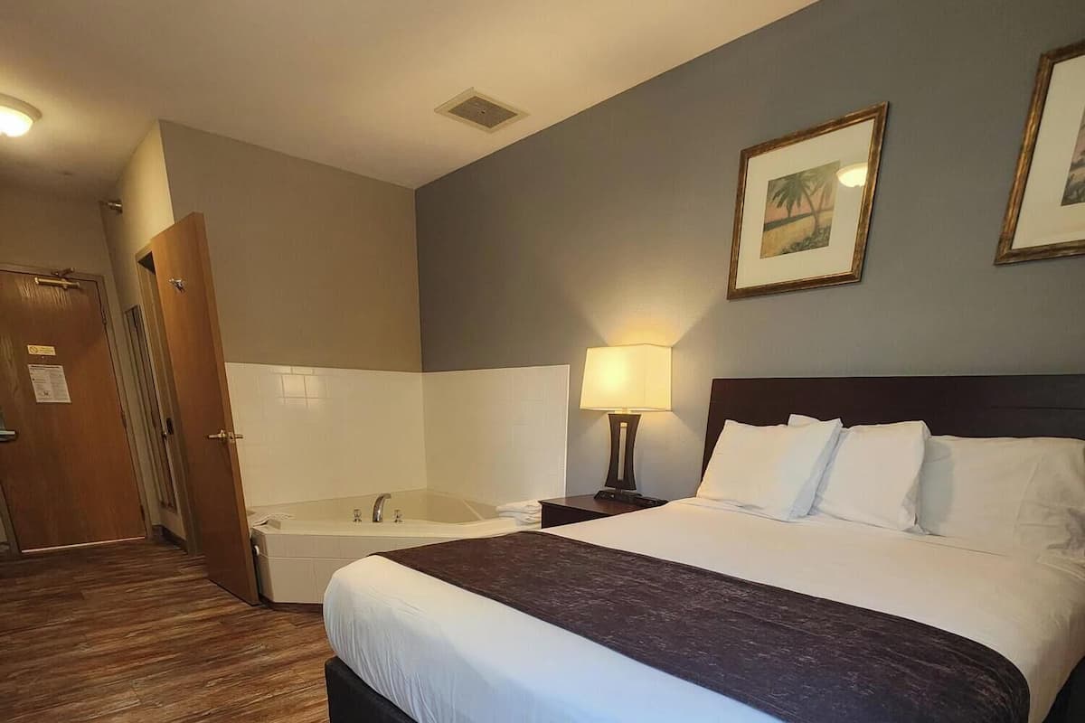 A hotel room with a large bed, a nightstand with a lamp, a bathroom with a bathtub, and wooden flooring.