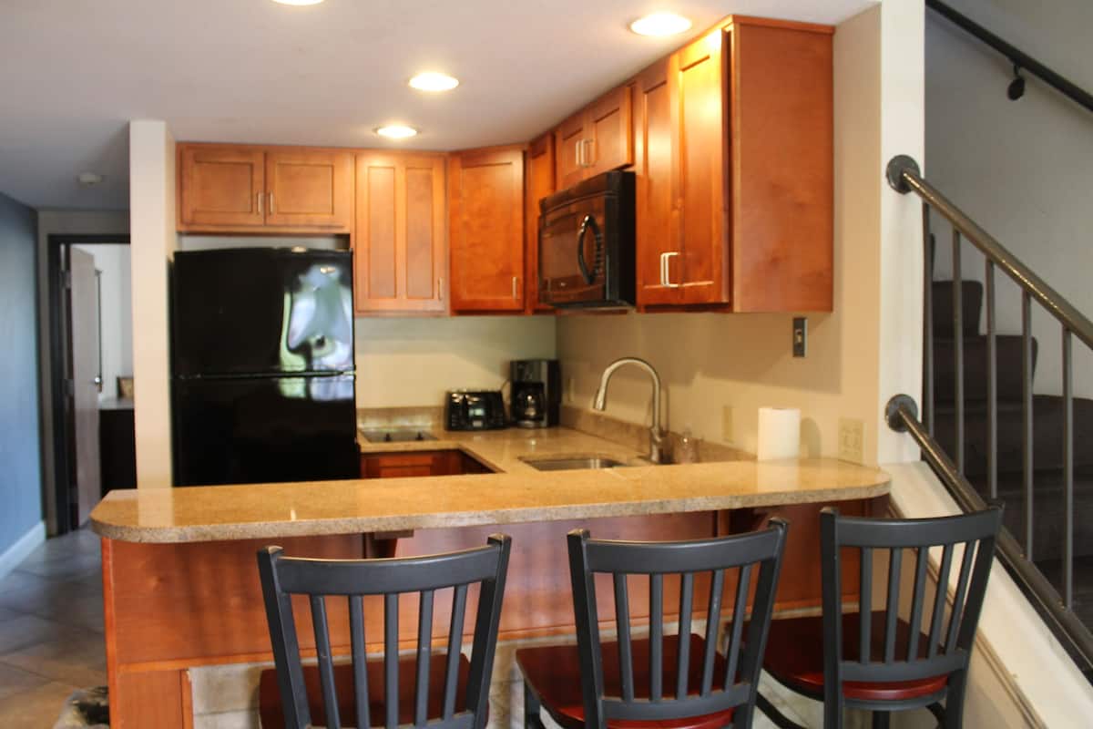 A kitchen with wooden cabinets, a black refrigerator, a microwave, and a granite countertop. There are three chairs around the island.