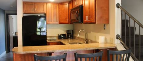 A kitchen with wooden cabinets, a black refrigerator, a microwave, and a granite countertop. There are three chairs around the island.