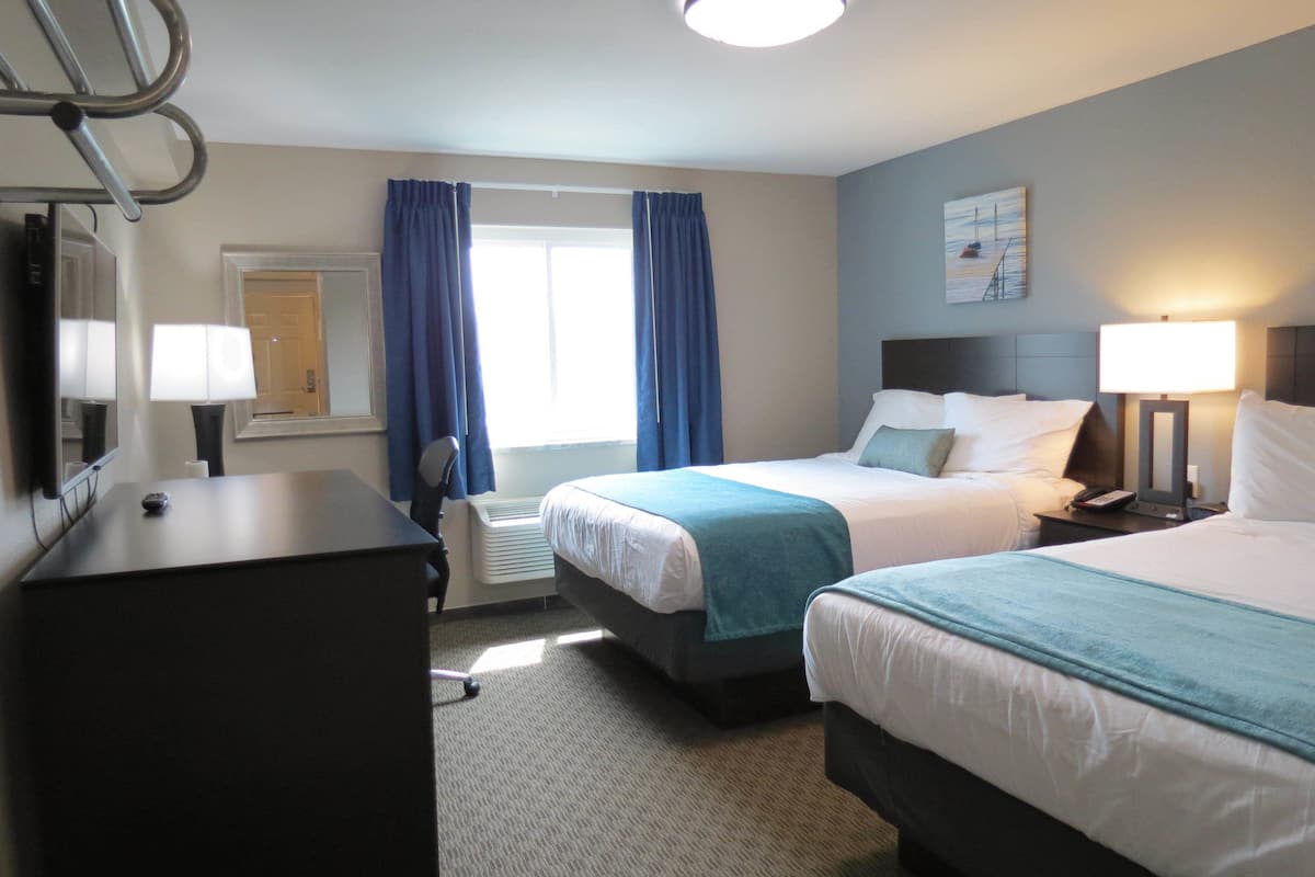 A hotel room with two beds, a desk, a chair, a desk lamp, a bedside table with a lamp, a mirror, and a window with curtains.