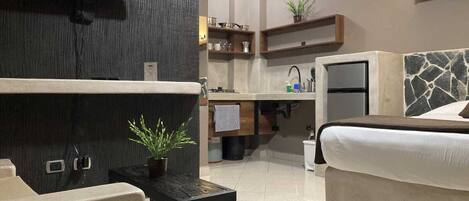 A modern hotel room with a bed, a small table with a plant, a TV mounted on the wall, a kitchenette, and a stone accent wall.