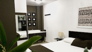 A modern hotel room with a large bed, a desk with a chair, a bathroom with a mirror and sink, and a wall-mounted lamp. - Ocean Breeze (Santo Domingo)
