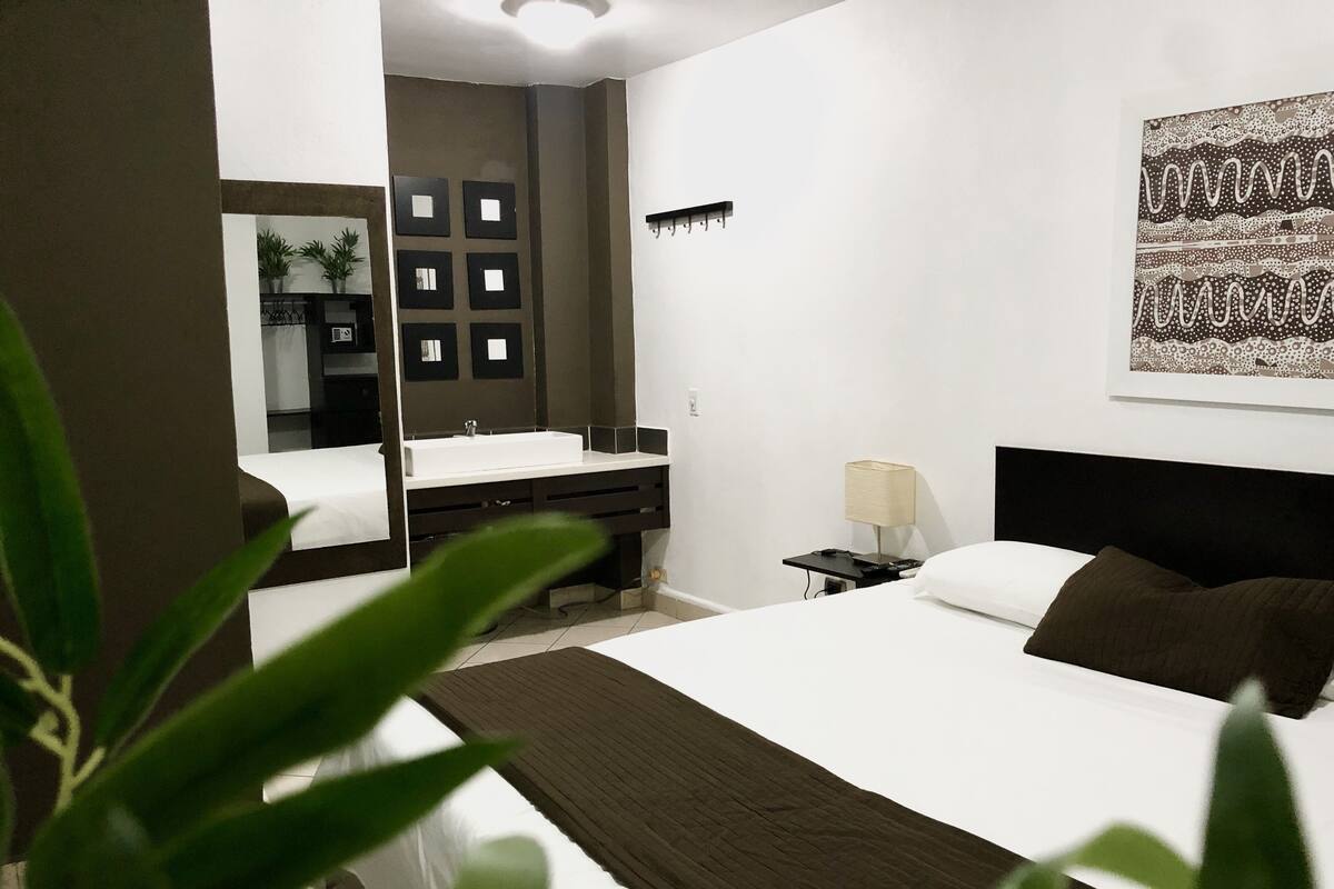 A modern hotel room with a large bed, a desk with a chair, a bathroom with a mirror and sink, and a wall-mounted lamp.