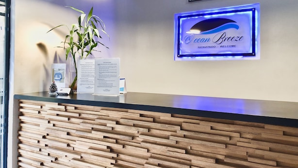A reception desk with a wooden panel design, a blue illuminated sign reading "Ocean Breeze," and a potted plant. - Ocean Breeze (Santo Domingo)