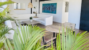 A modern, open-plan living area with a white sofa, a wooden coffee table, and a round table. There are potted plants, a painting on the wall, and a ceiling with exposed beams. - Ocean Breeze (Santo Domingo)