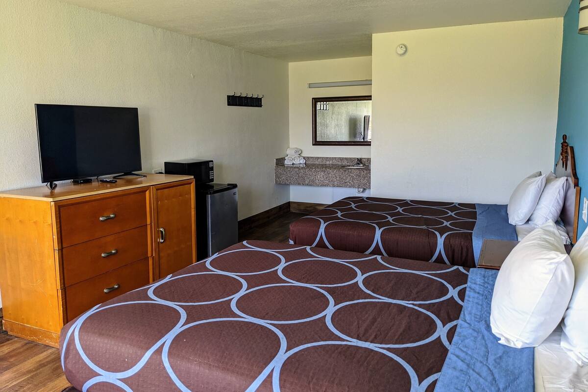 A bedroom with a bed, a television on a wooden dresser, a small kitchenette, and a bathroom with a mirror and sink.